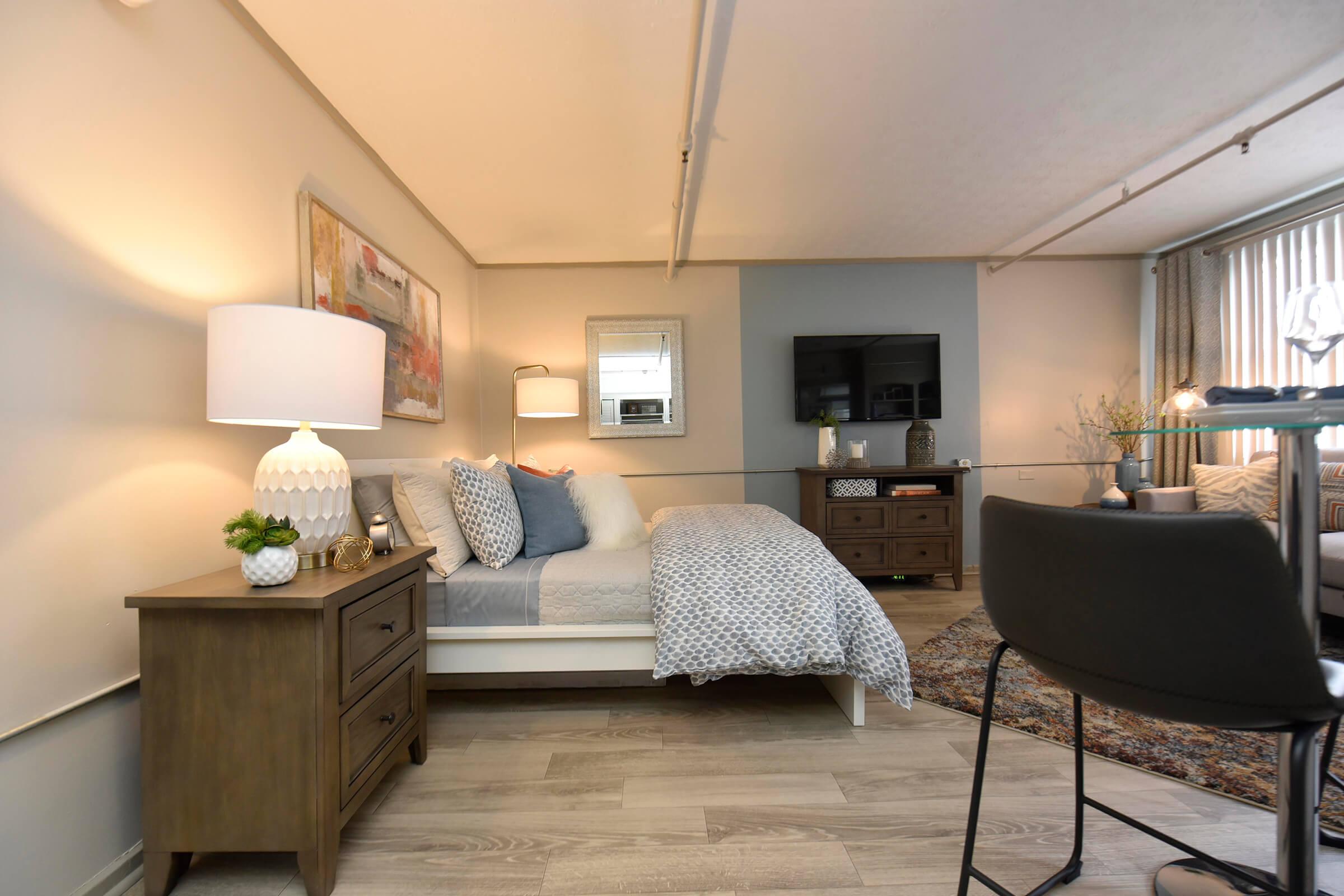 A cozy living space featuring a neatly made bed with decorative pillows, a bedside lamp on a wooden nightstand, a television mounted on the wall, and a comfortable seating area with a rug. Natural light streams in, highlighting the modern decor and inviting atmosphere.