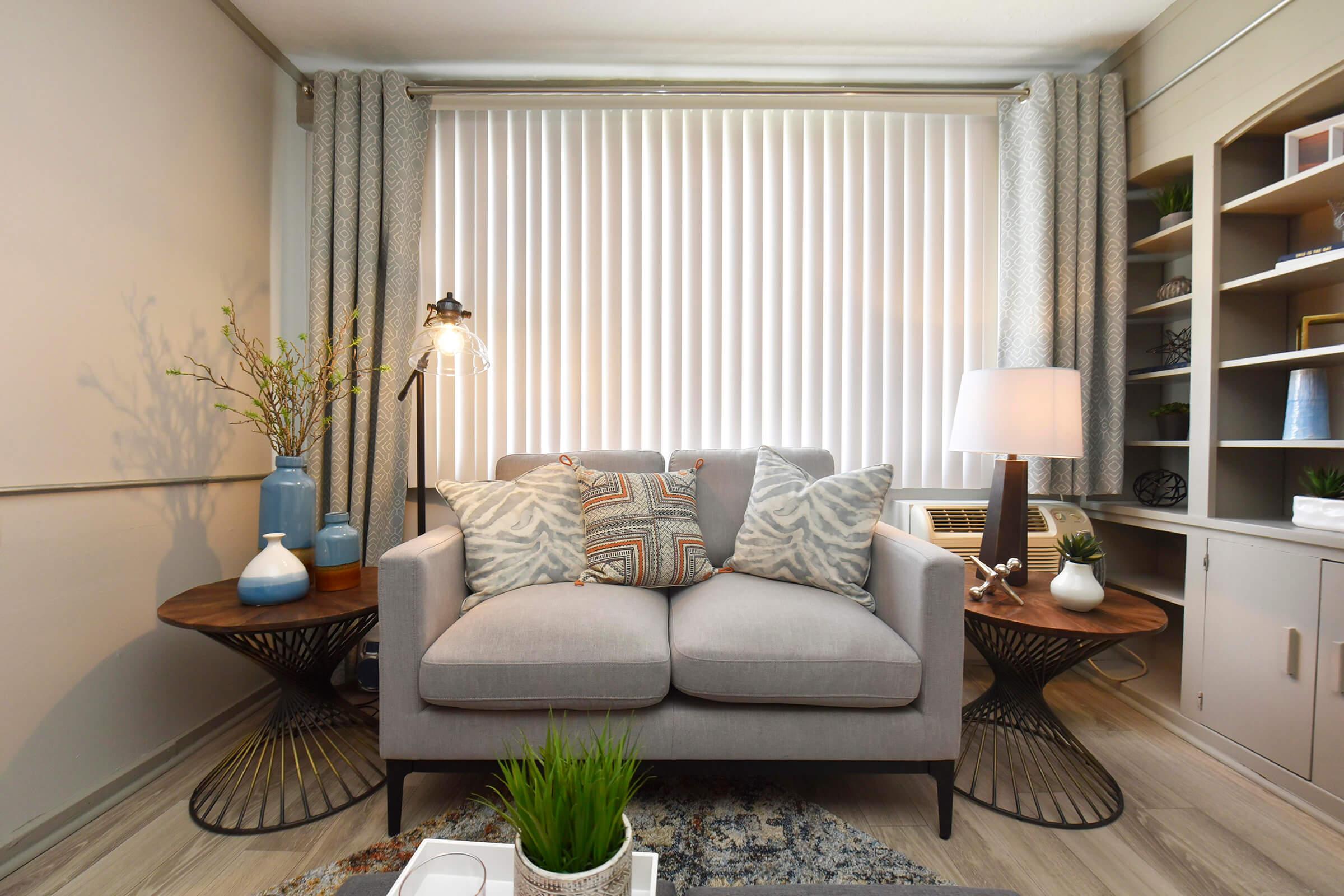 A cozy living room setup featuring a gray sofa with decorative pillows, two round side tables, a lamp, and potted plants. Light-colored curtains cover a window, and there are decorative vases on the side tables and shelves. The warm ambiance is enhanced by natural light and modern decor elements.