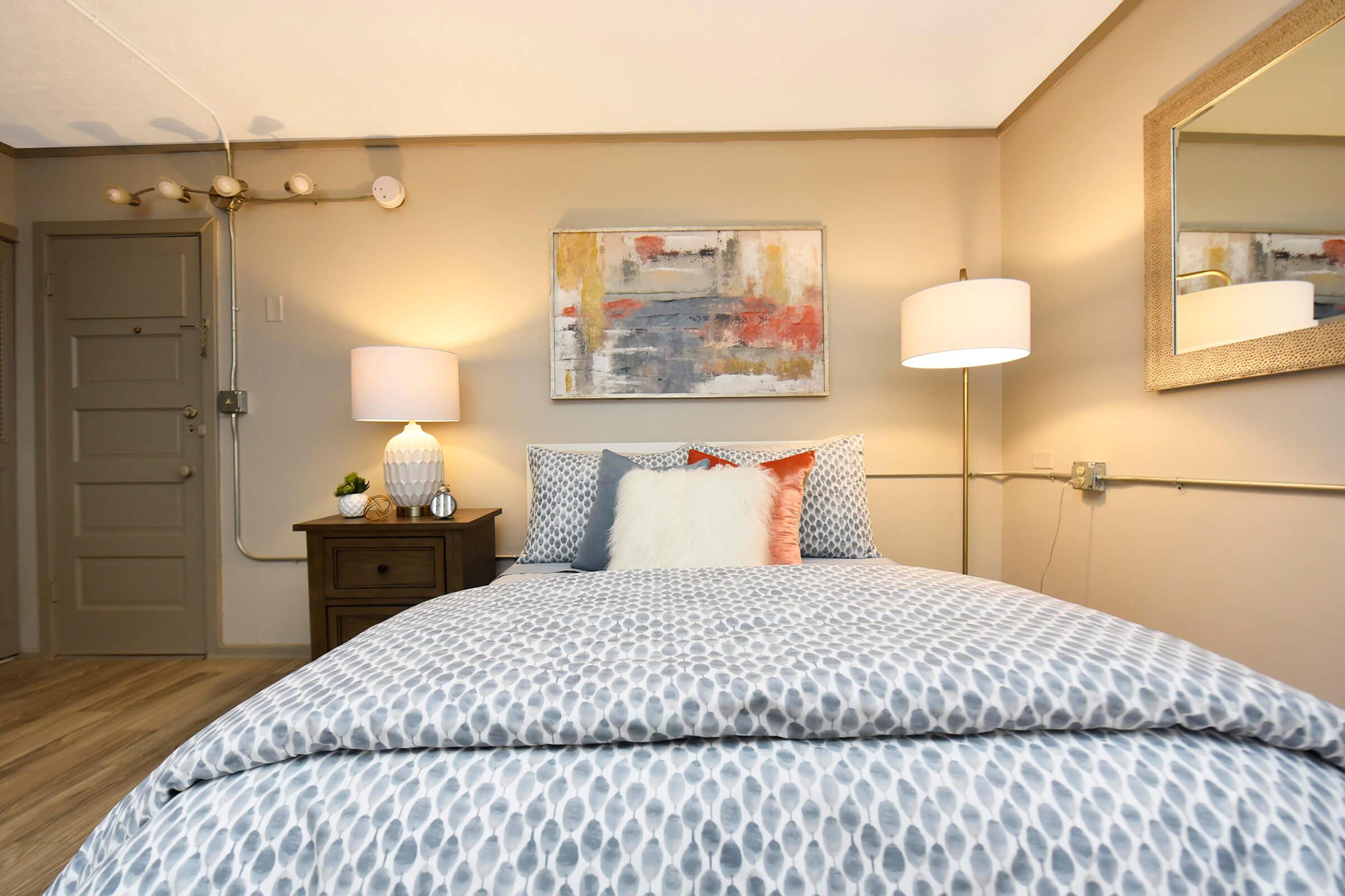 A cozy bedroom featuring a queen-sized bed with patterned bedding, two bedside lamps, and a decorative pillow. A piece of abstract art hangs on the wall above the bed, complemented by a mirror. The warm color palette and wooden nightstand create a welcoming atmosphere.