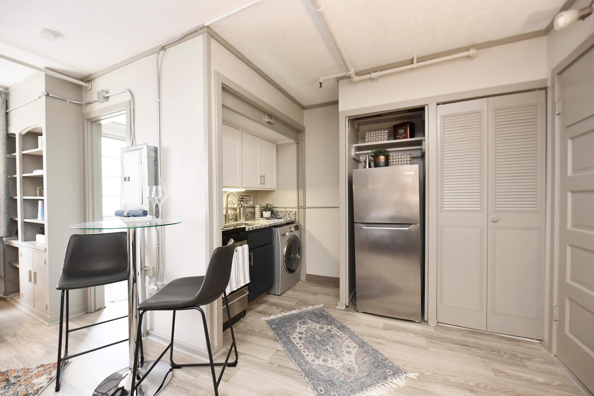 A modern kitchen featuring a stainless steel refrigerator, washing machine, and white cabinetry. There is a small dining area with two black bar stools and a glass table. Shelving is displayed on the wall, and a patterned rug is on the floor. The space has a clean, minimalist design with gray accents.