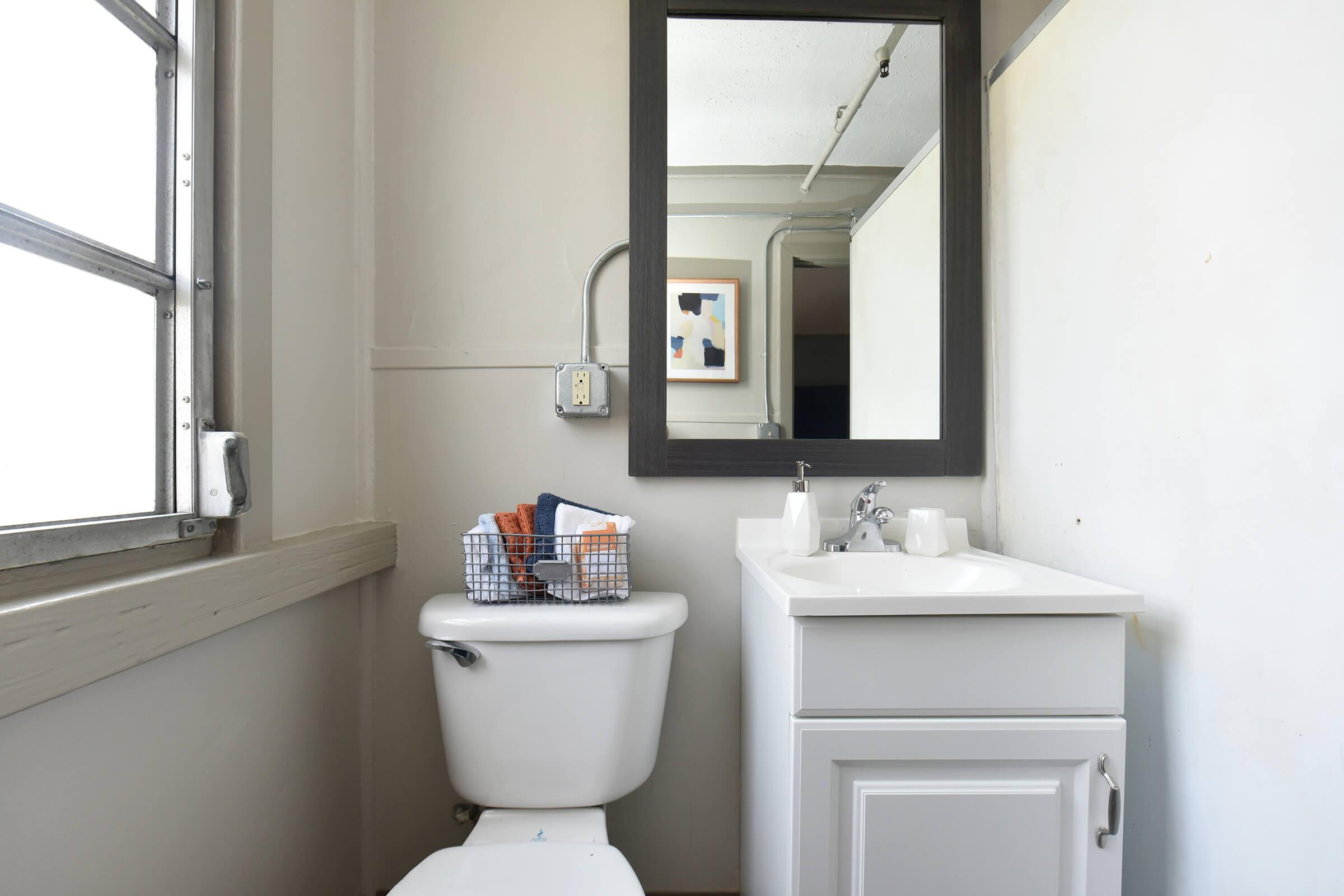 A small bathroom featuring a white toilet and a compact sink with a cabinet underneath. Above the sink, there's a rectangular mirror in a dark frame. A wire basket holding towels is placed on the sink, and the walls are painted a light color, creating a clean and minimalistic look.