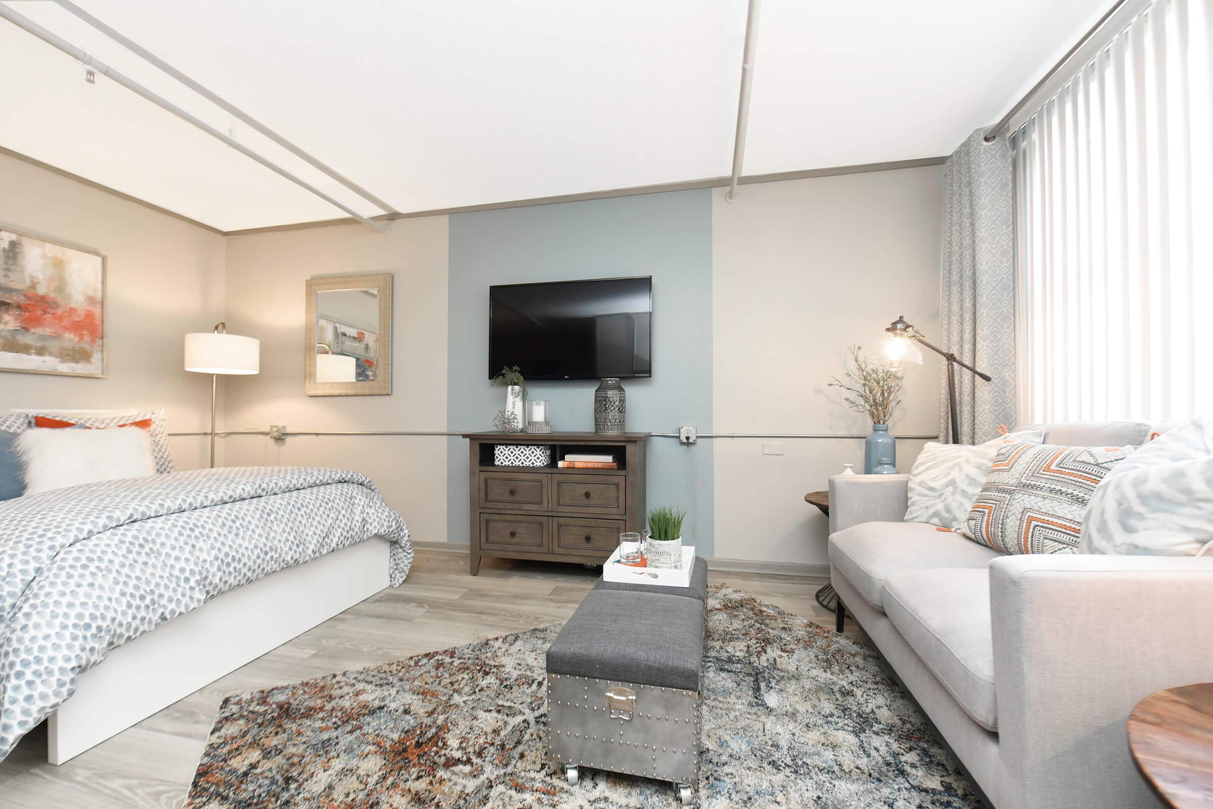 A cozy bedroom featuring a queen-sized bed with a patterned duvet, a modern gray sofa adorned with decorative pillows, a coffee table, and a large TV mounted on the wall. Natural light streams through window treatments, and decorative elements like plants and artwork enhance the inviting atmosphere.