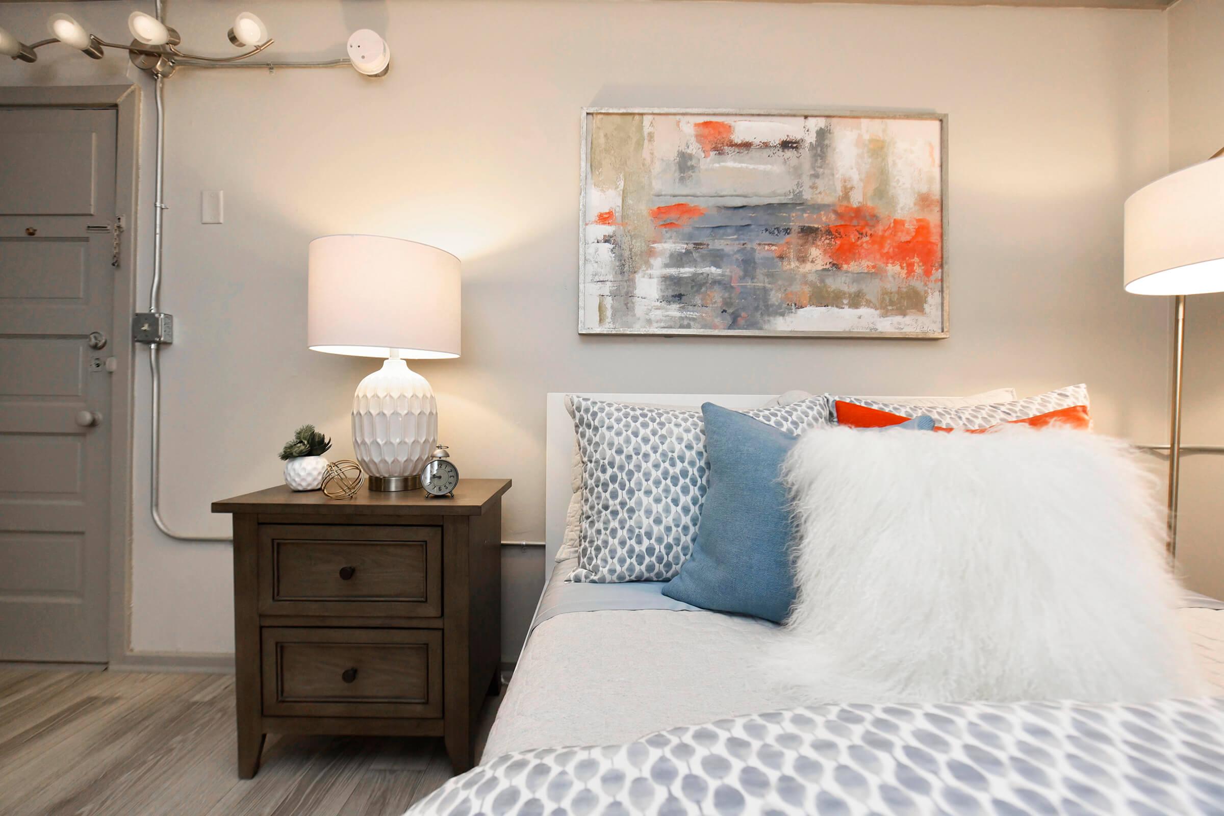 A cozy bedroom featuring a bed with decorative pillows, a wooden nightstand with a small plant and decorative items, and two table lamps. The wall above the bed showcases an abstract painting with shades of gray, orange, and white. Soft lighting creates a warm, inviting atmosphere.