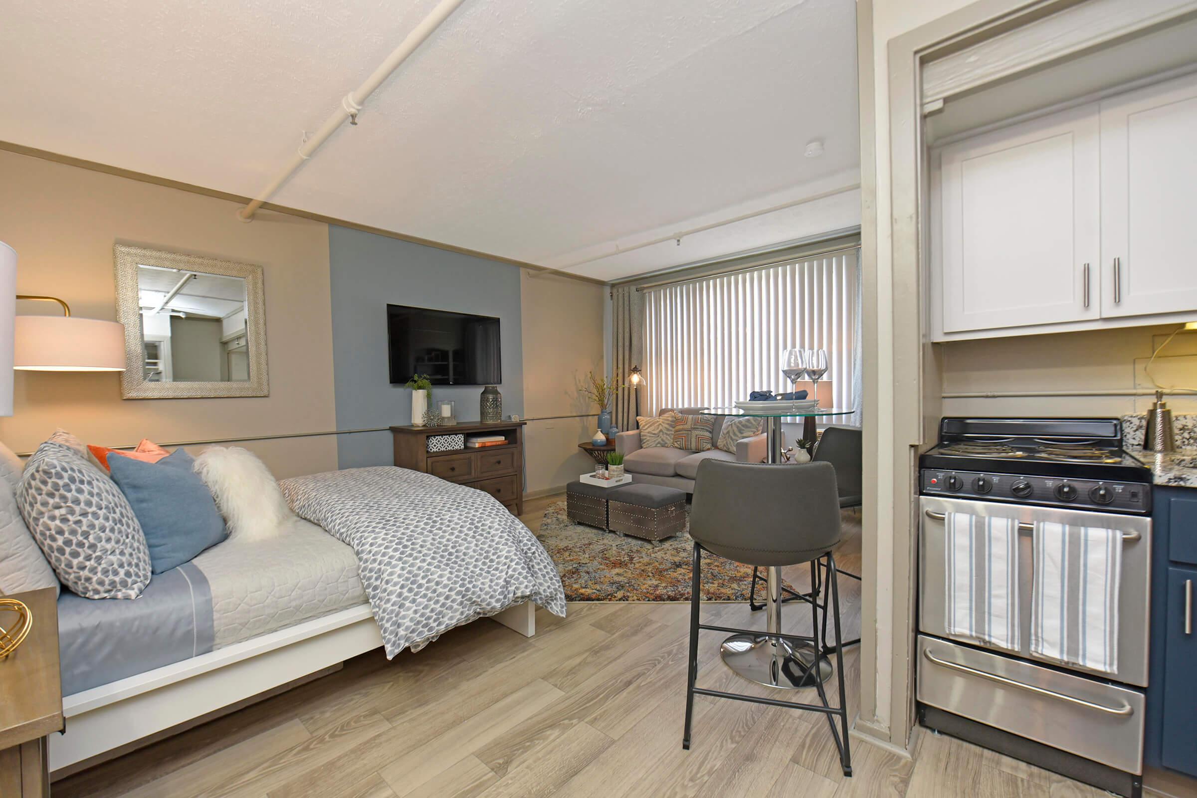 A modern studio apartment featuring a bed with patterned bedding, a cozy living area with a gray couch, a flat-screen TV on the wall, and a small kitchenette with a stove. Natural light enters through large windows with blinds, creating a warm and inviting atmosphere.