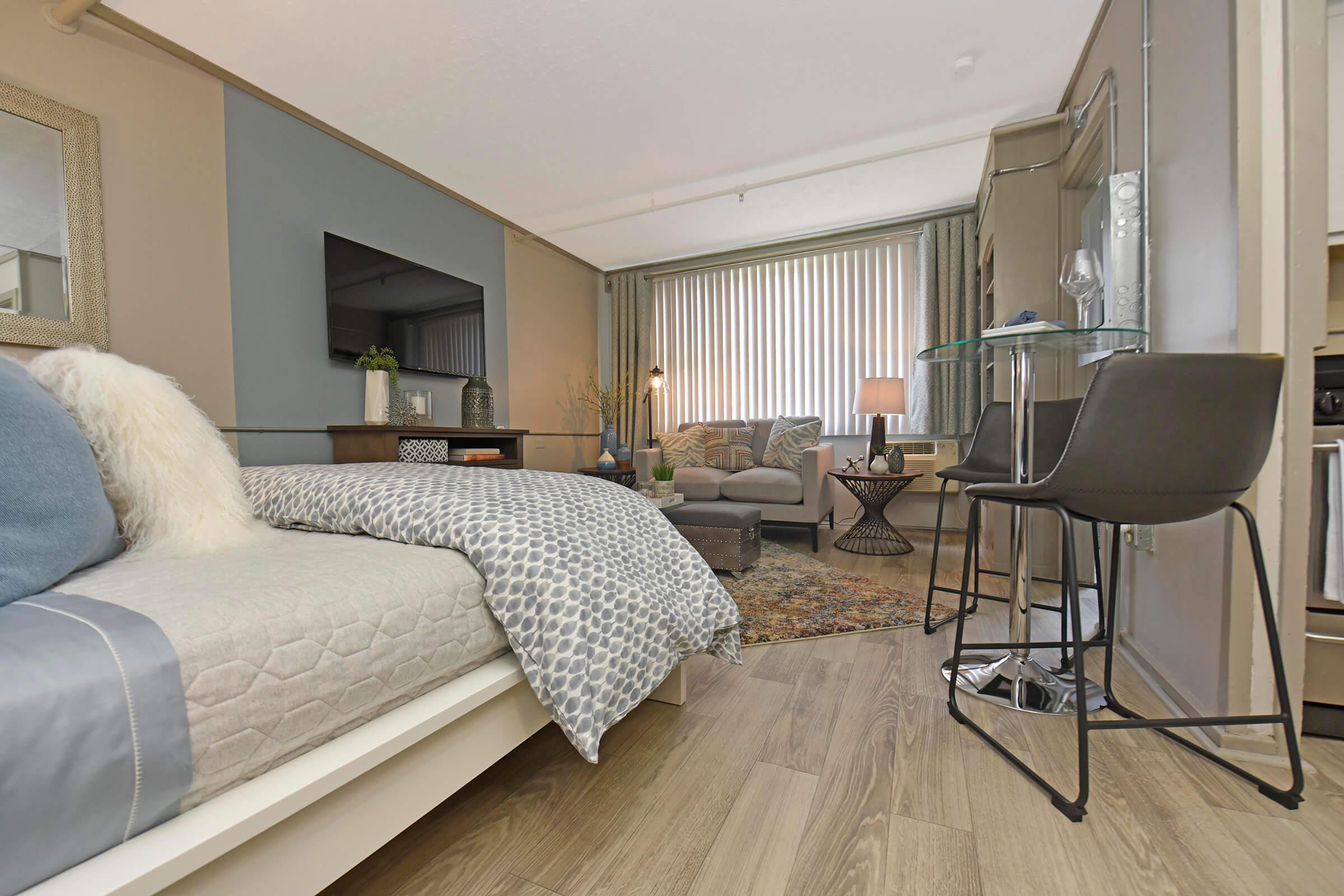 A modern and stylish studio apartment featuring a bed with decorative pillows, a cozy seating area with a sofa and lamp, a small dining table with a chair, and a television mounted on the wall. Large windows with light curtains allow natural light to fill the room, complemented by wood-like flooring.