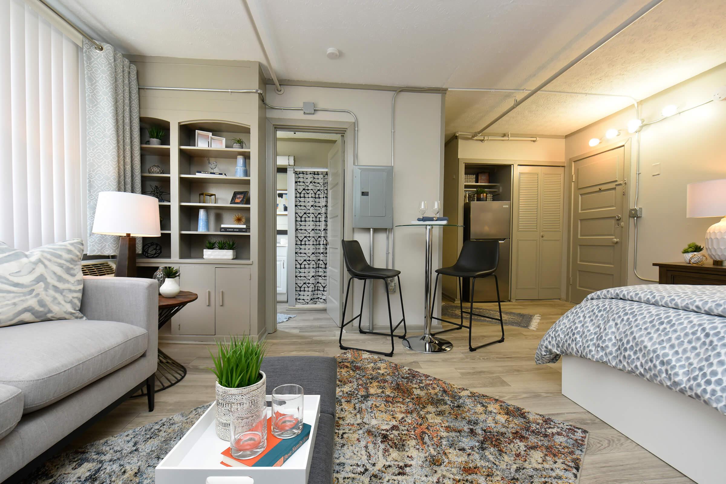 A cozy studio apartment featuring a small living area with a gray sofa and decorative pillows, a side table with drinks, a dining space with two black chairs, and a kitchenette in the background. The walls are light-colored, and there's a rug on the floor, creating a welcoming atmosphere.