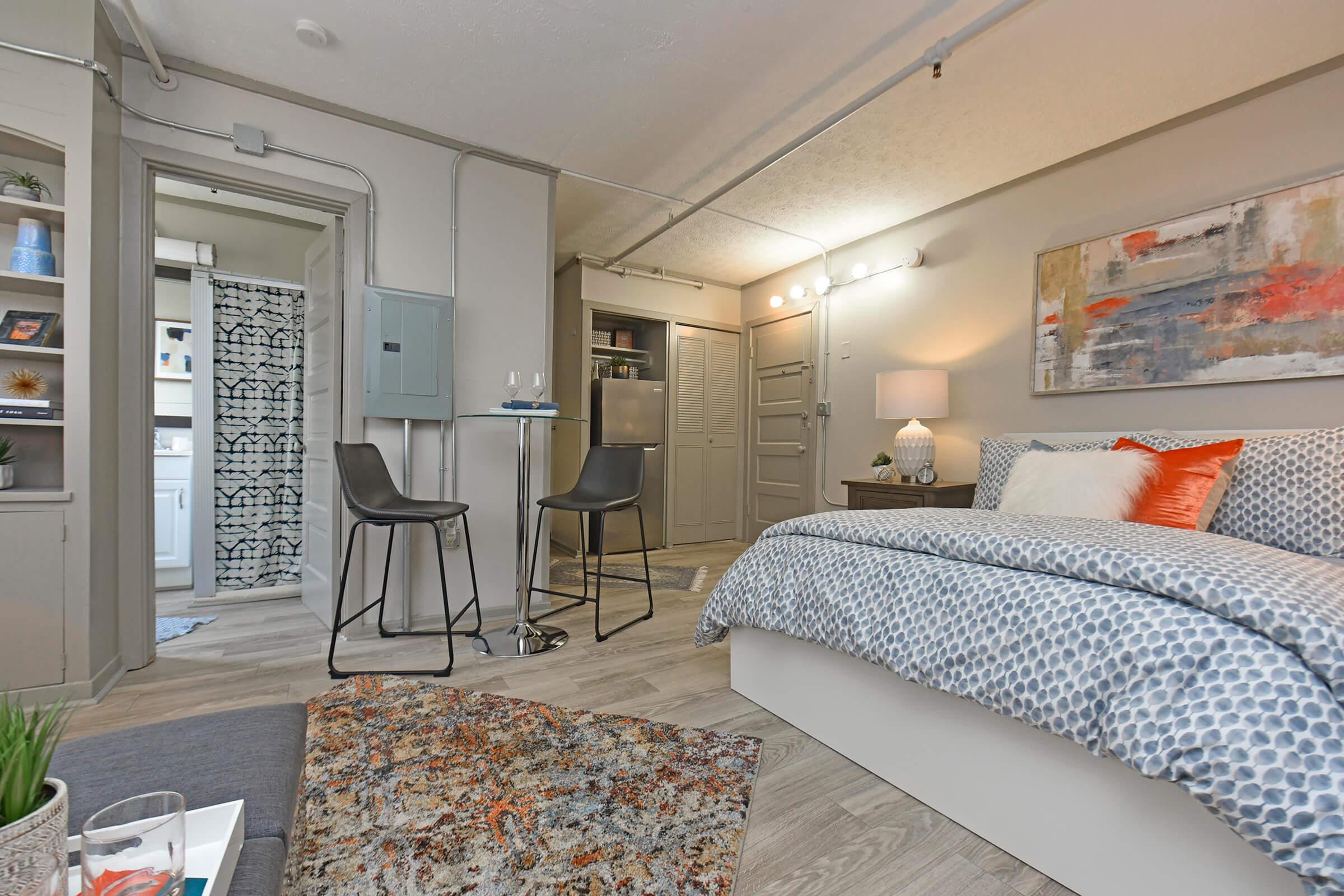 A cozy studio apartment featuring a bed with patterned bedding, a small dining table with two chairs, and a decorative rug. The space includes a kitchenette area and a bathroom visible through an open door. Soft lighting enhances the warm and inviting atmosphere.