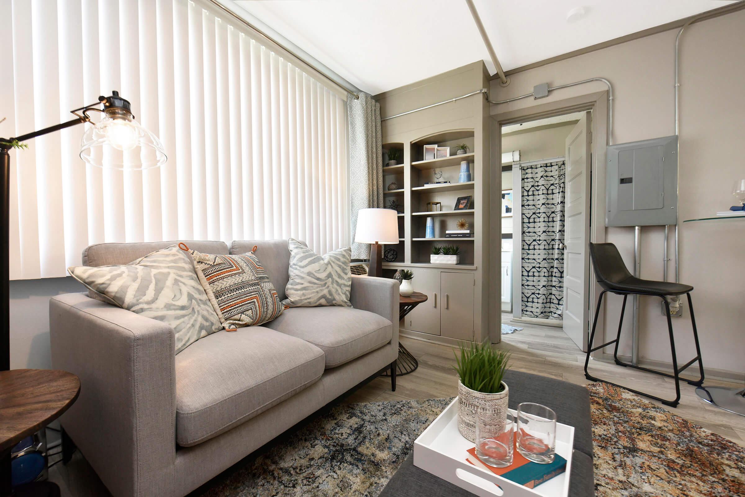 A cozy living room featuring a light gray sofa with patterned throw pillows, a wooden side table, and a decorative rug. In the background, there is a lamp, a bookshelf filled with items, and a doorway leading to another space. The room has natural light from large blinds and a modern design aesthetic.