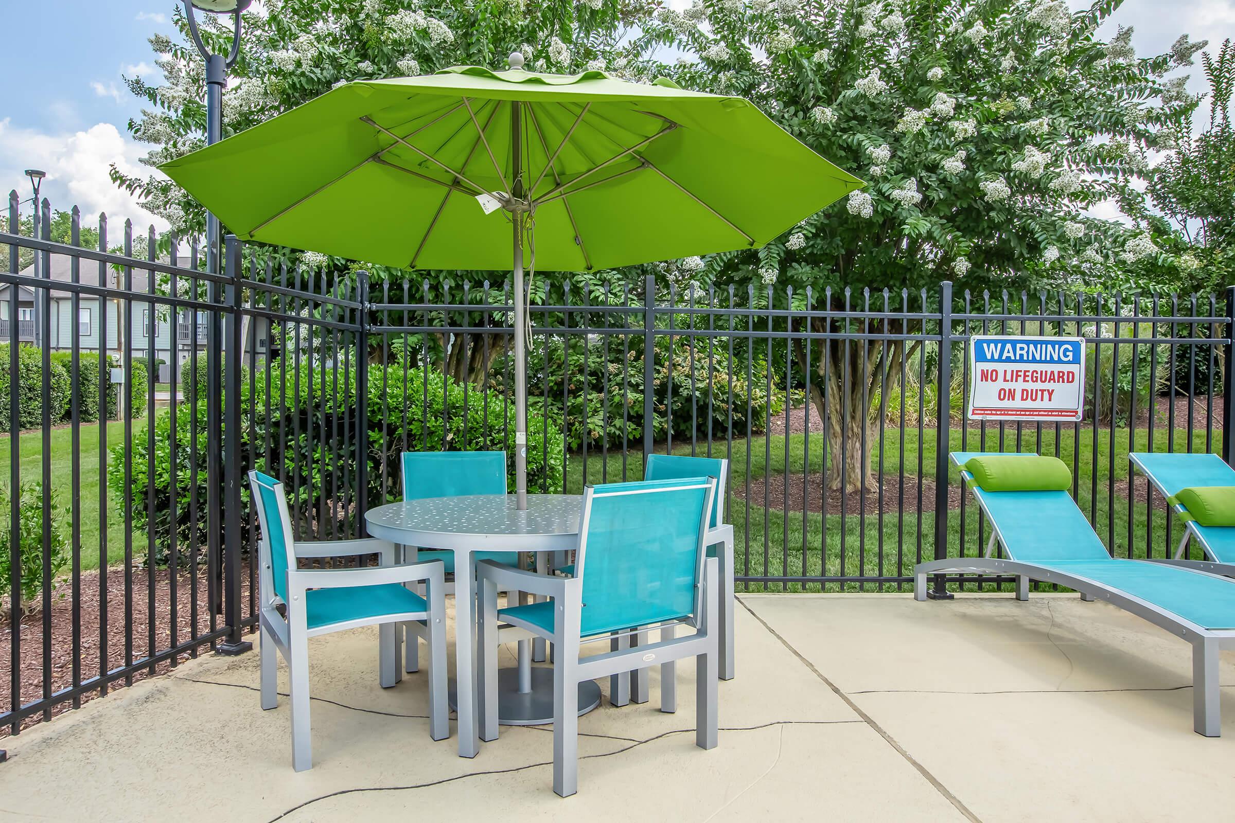 A poolside area featuring a round table with four turquoise chairs and a large green umbrella. To the side, there are two lounge chairs and a warning sign indicating "No Lifeguard on Duty." The background includes green shrubs and trees, enclosed by a black fence.