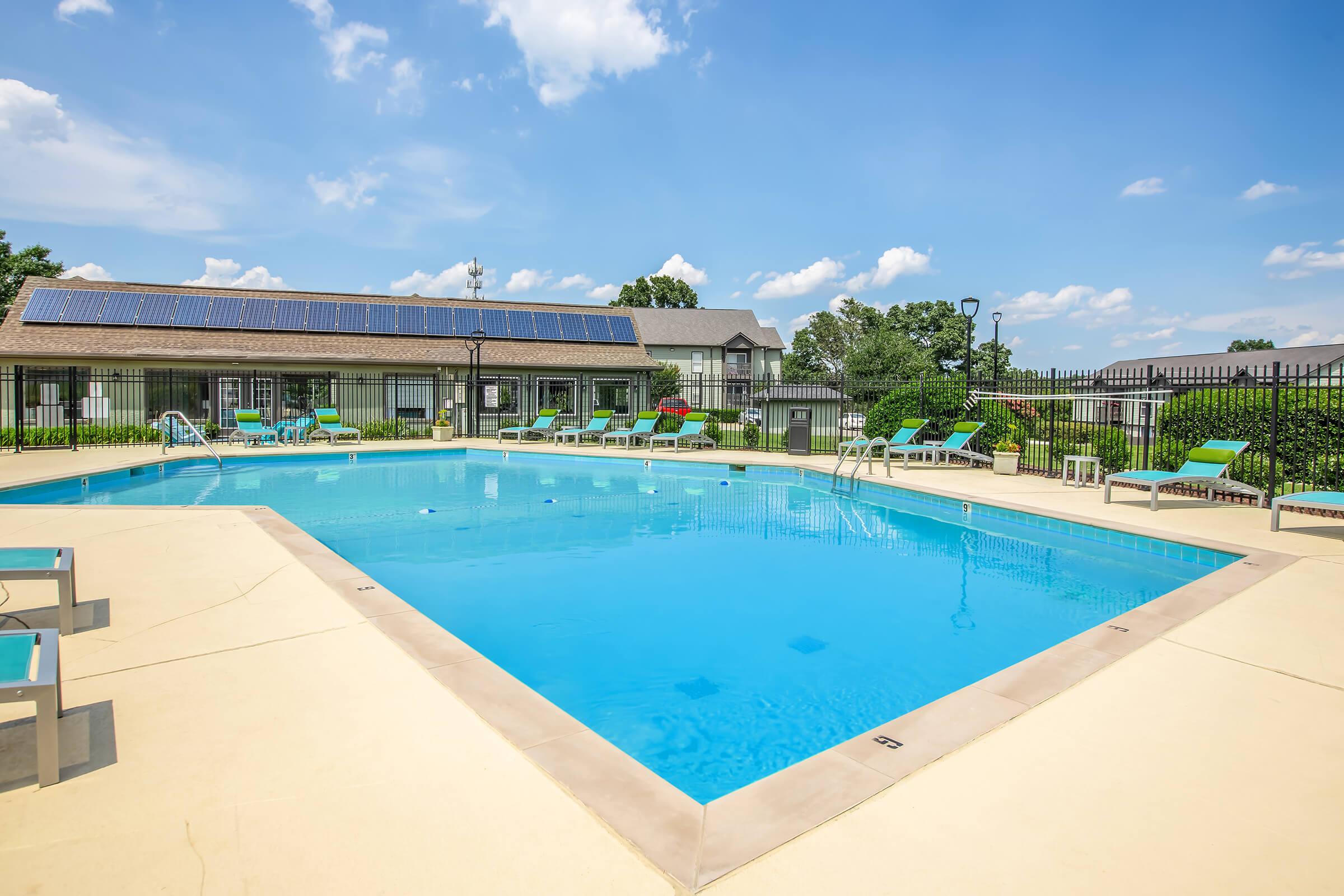 A bright blue swimming pool with clear water, surrounded by lounge chairs. There are green bushes and trees in the background, a building with solar panels near the pool, and a clear blue sky with scattered clouds. The area is well-maintained and inviting for relaxation.