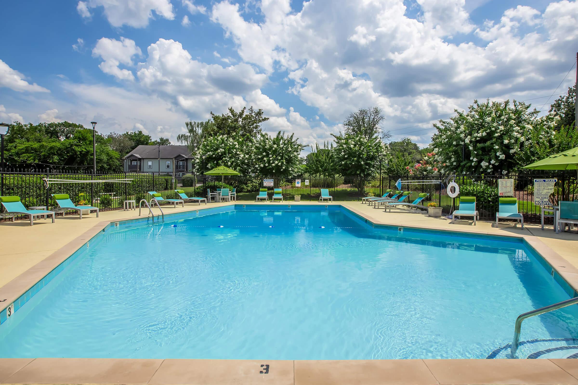 A clear blue swimming pool surrounded by lounge chairs under green umbrellas. Lush trees and flowering bushes line the pool area, with partly cloudy skies overhead. The setting is inviting and serene, ideal for relaxation and recreation.