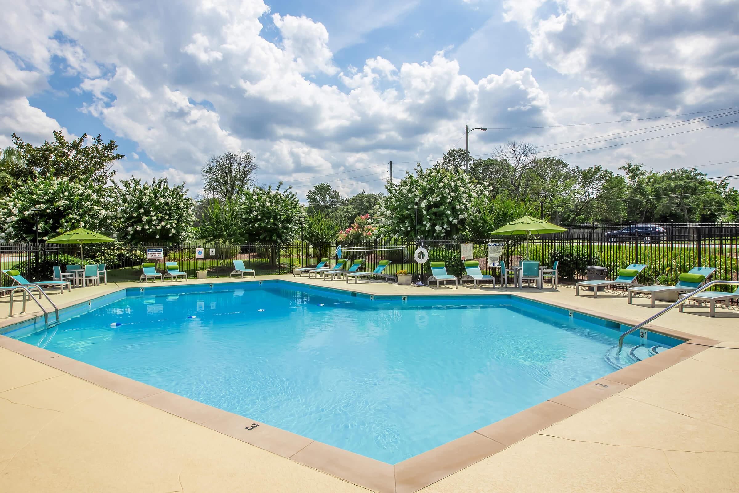 A clear blue swimming pool surrounded by lounge chairs and green umbrellas. Lush greenery and flowering bushes create a serene atmosphere, with a cloudy sky above. The pool area is well-maintained, inviting for relaxation and leisure.