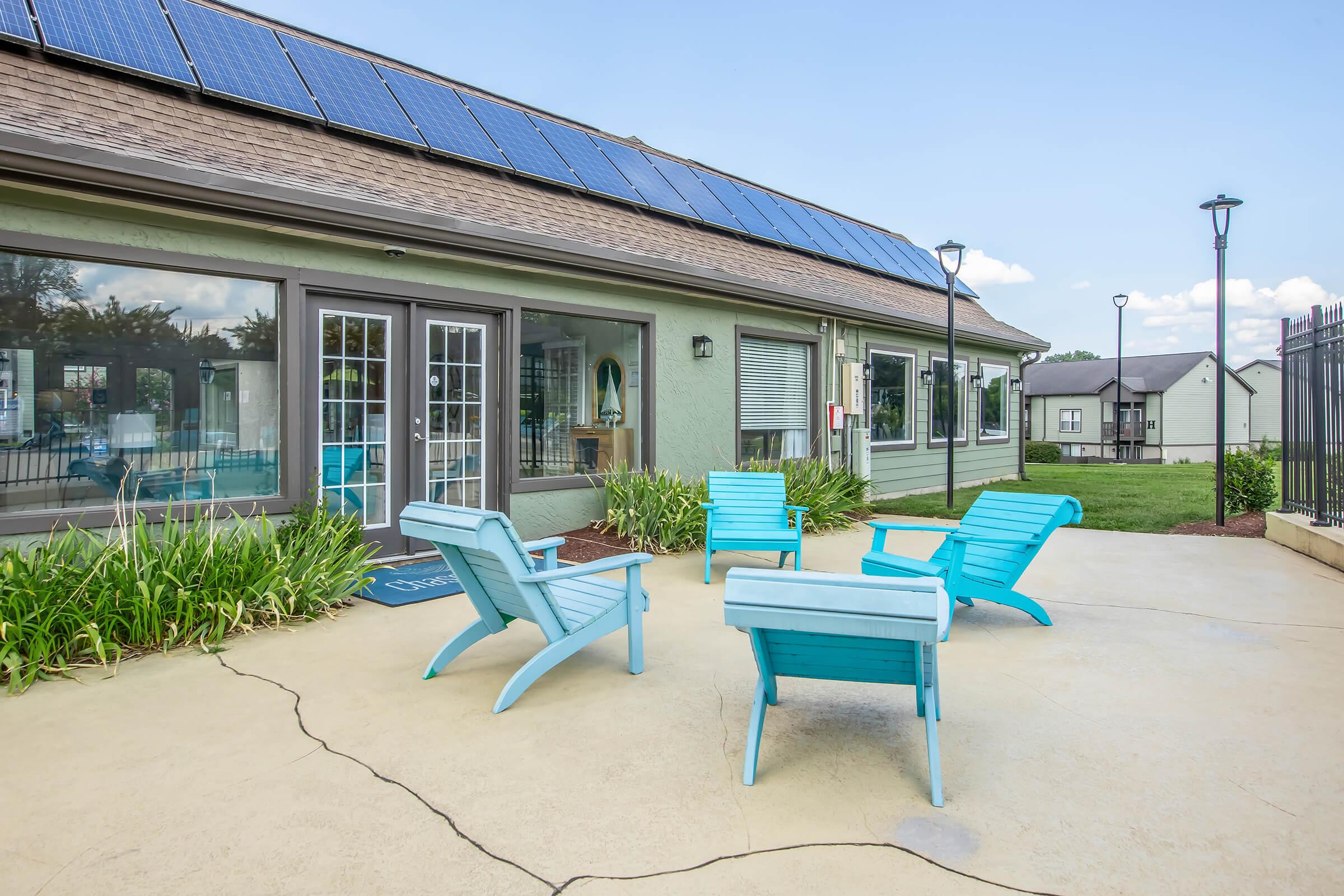 A patio area featuring several light blue outdoor chairs arranged around a small table. In the background, there is a building with large windows and solar panels on the roof, surrounded by green grass and landscaping. The sky is clear with a few clouds, creating a pleasant outdoor setting.