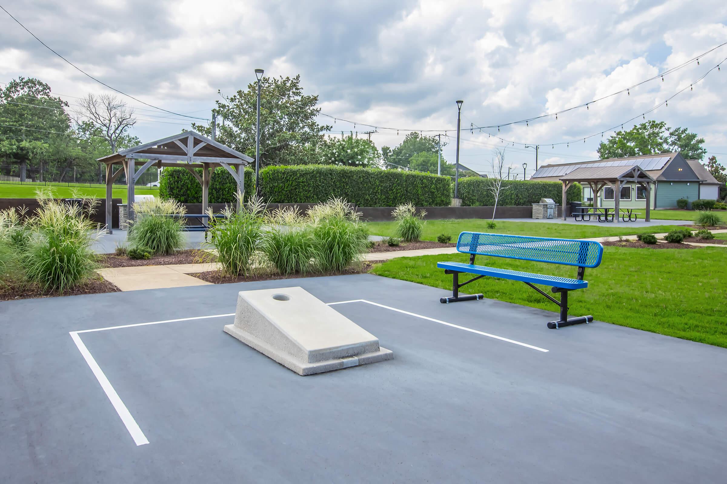 A view of a well-maintained outdoor area featuring a cornhole game setup with a beanbag toss platform, a blue bench, and greenery. There are two gazebos in the background and string lights above, creating a relaxing atmosphere in a park-like setting.
