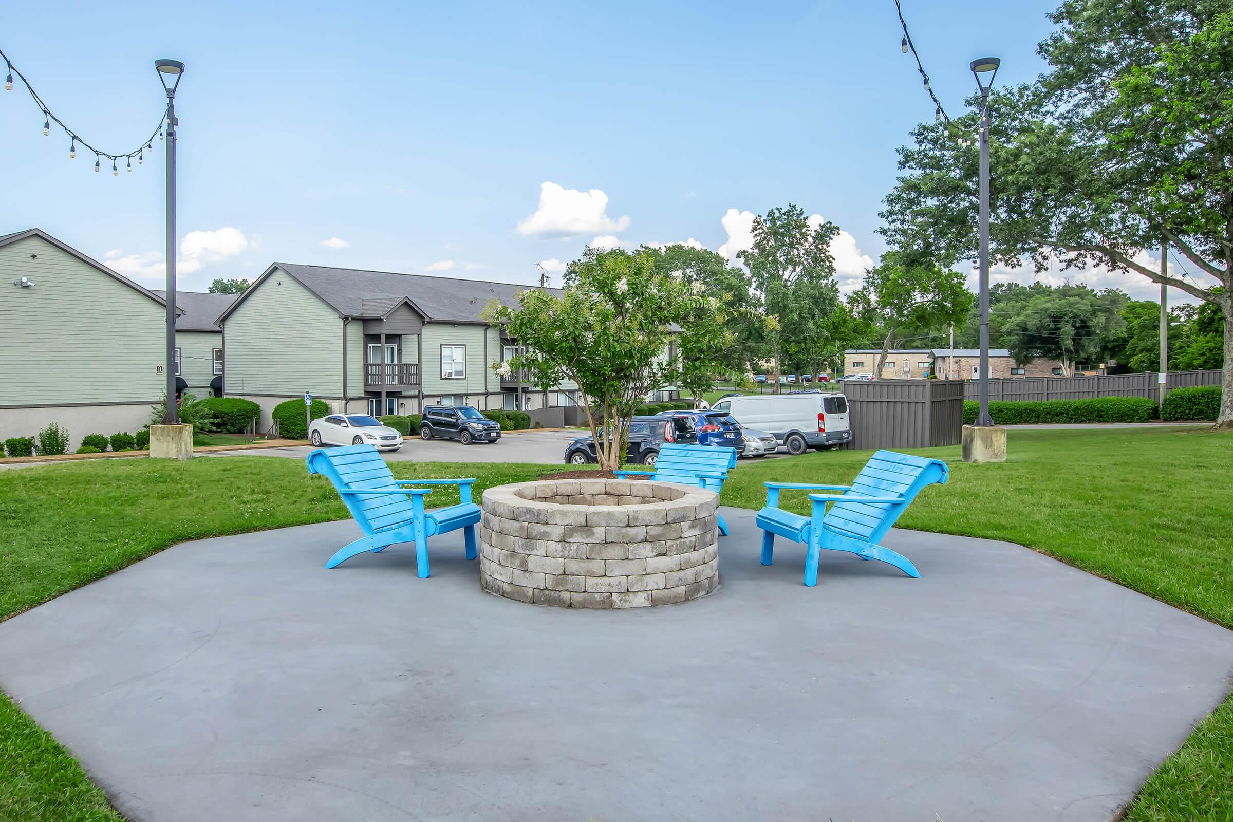 A cozy outdoor seating area with four bright blue Adirondack chairs arranged around a circular stone fire pit. Lush green grass surrounds the space, and a small tree is planted in the center. In the background, there are residential buildings and parked cars, under a clear blue sky with scattered clouds.