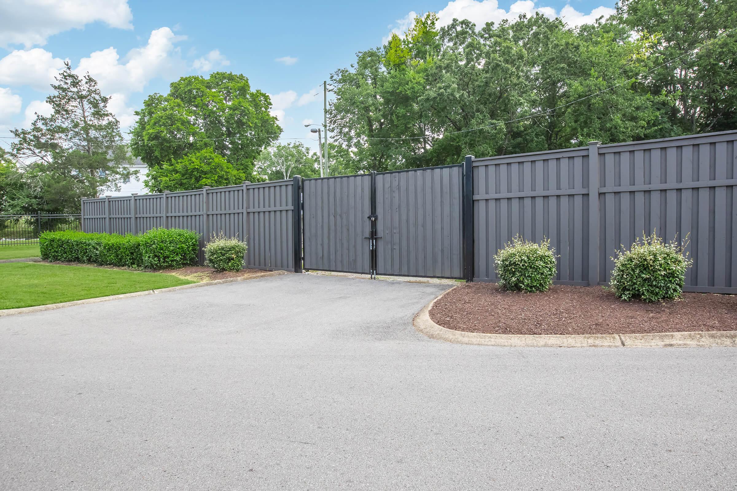 A modern gray gated entryway surrounded by lush green trees and shrubs, featuring a paved driveway leading up to the closed gate, set against a partly cloudy sky.