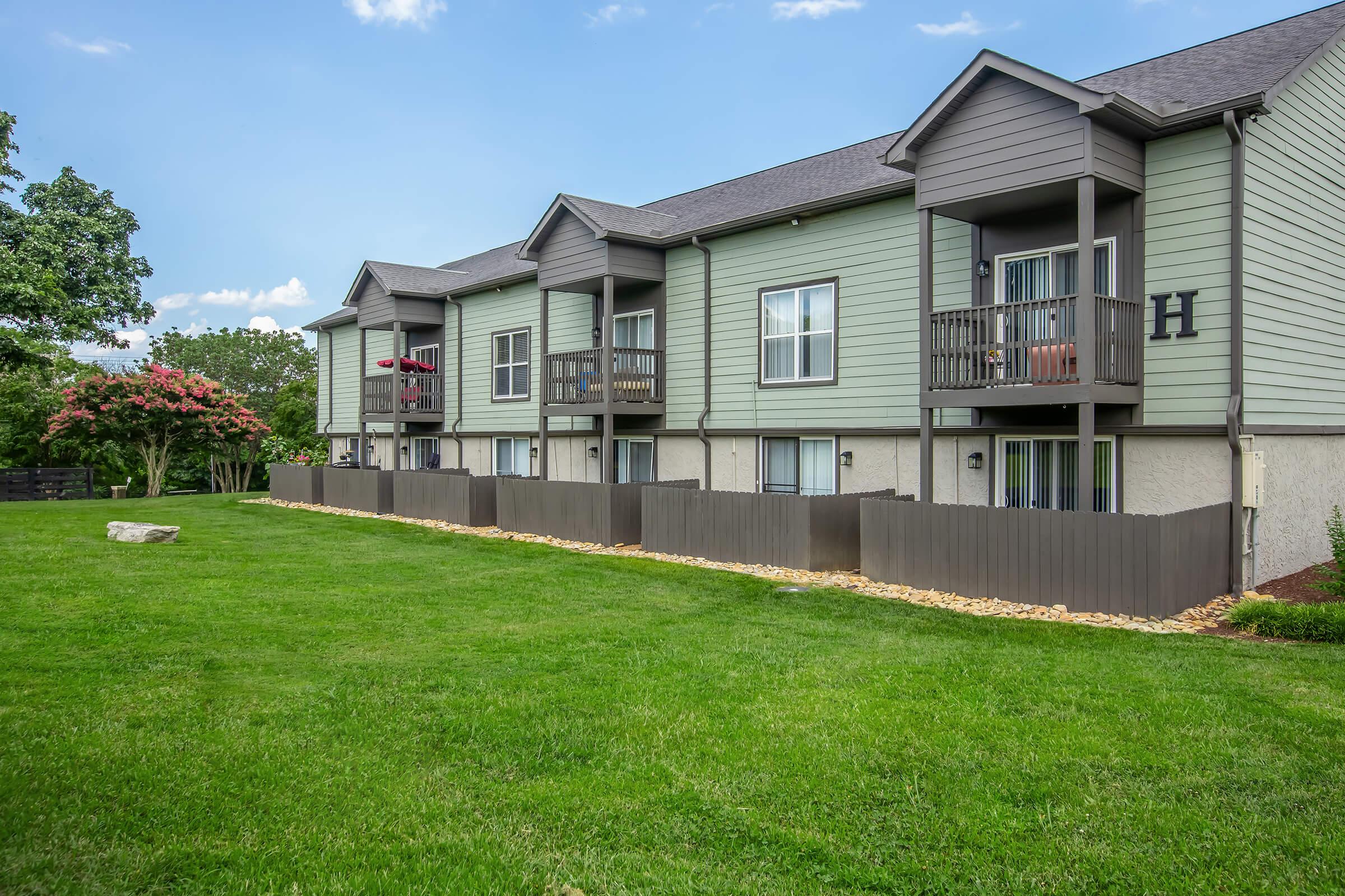 A row of green, two-story apartment buildings with balconies, surrounded by a well-maintained grassy area. There are decorative rocks along the bottom of the fences, and trees in the background against a clear blue sky. The setting appears inviting and well-kept.