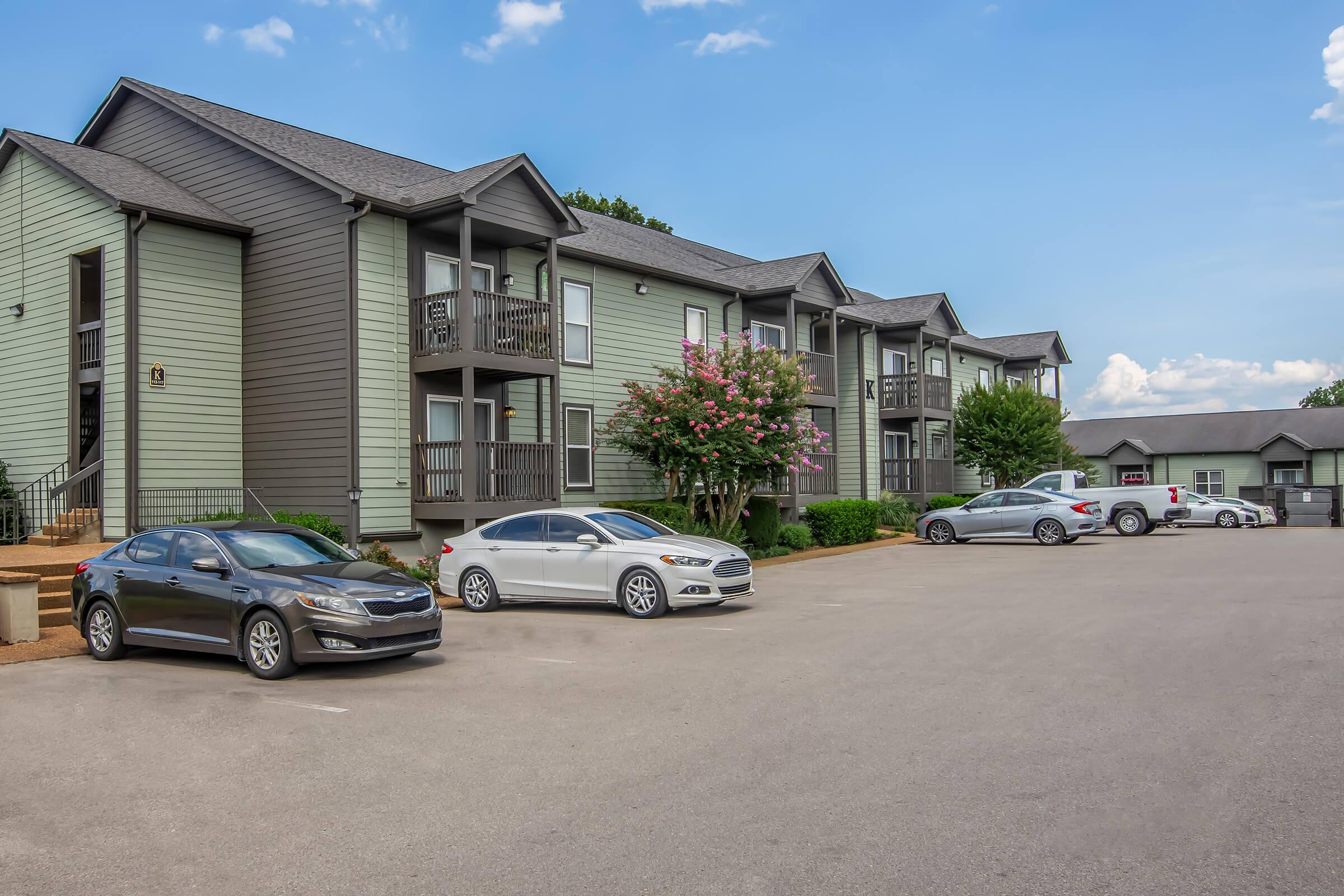 A row of two-story apartment buildings with a light green exterior. Several cars are parked in the foreground, including a black sedan, a white sedan, and a few other vehicles. The scene is set under a bright blue sky with a few clouds, and there are trees and flowering plants nearby.