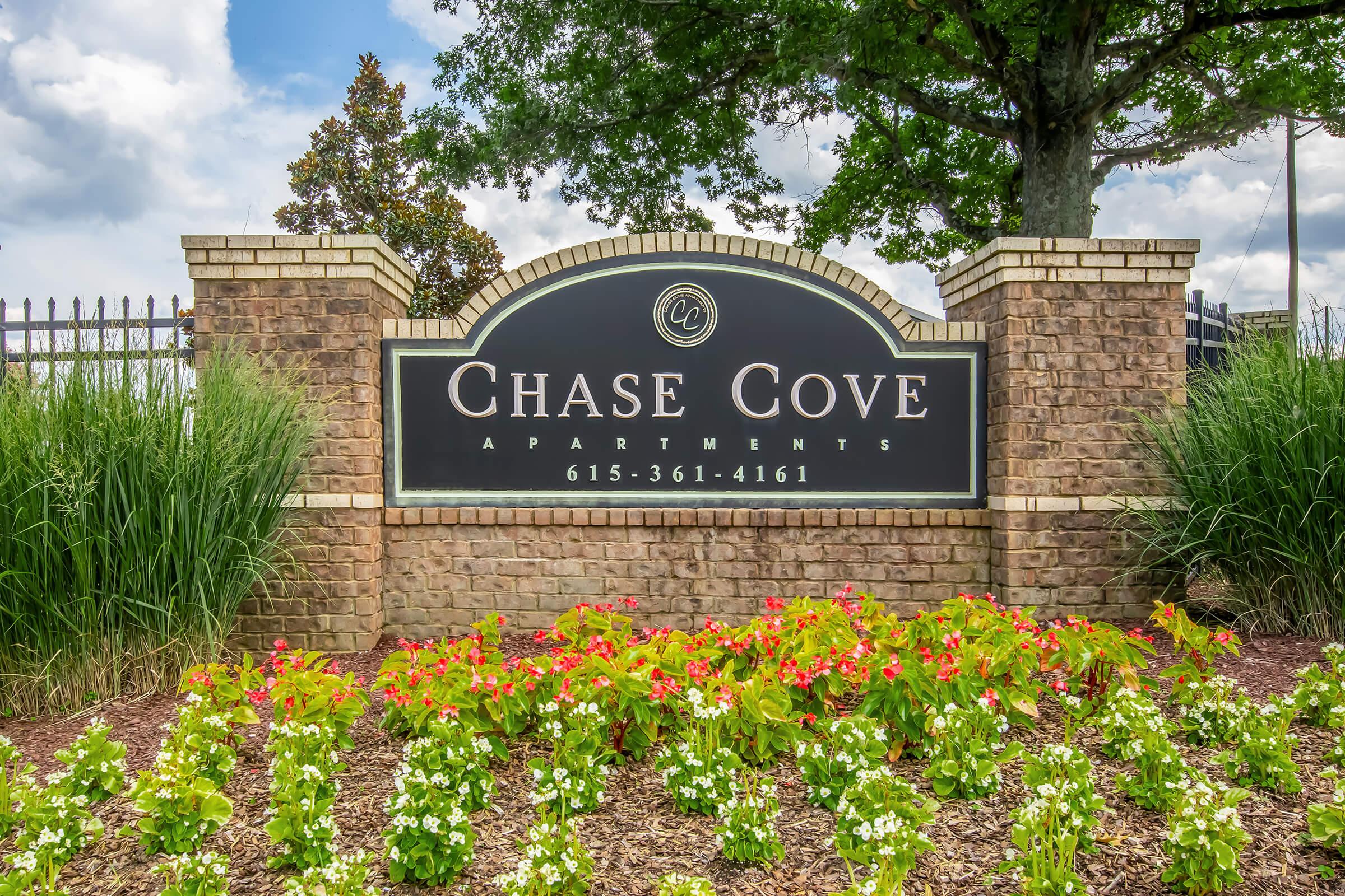 Sign for Chase Cove Apartments with a black background and white text, surrounded by colorful flower beds and greenery, under a cloudy sky. The sign includes the phone number 615-361-4161.