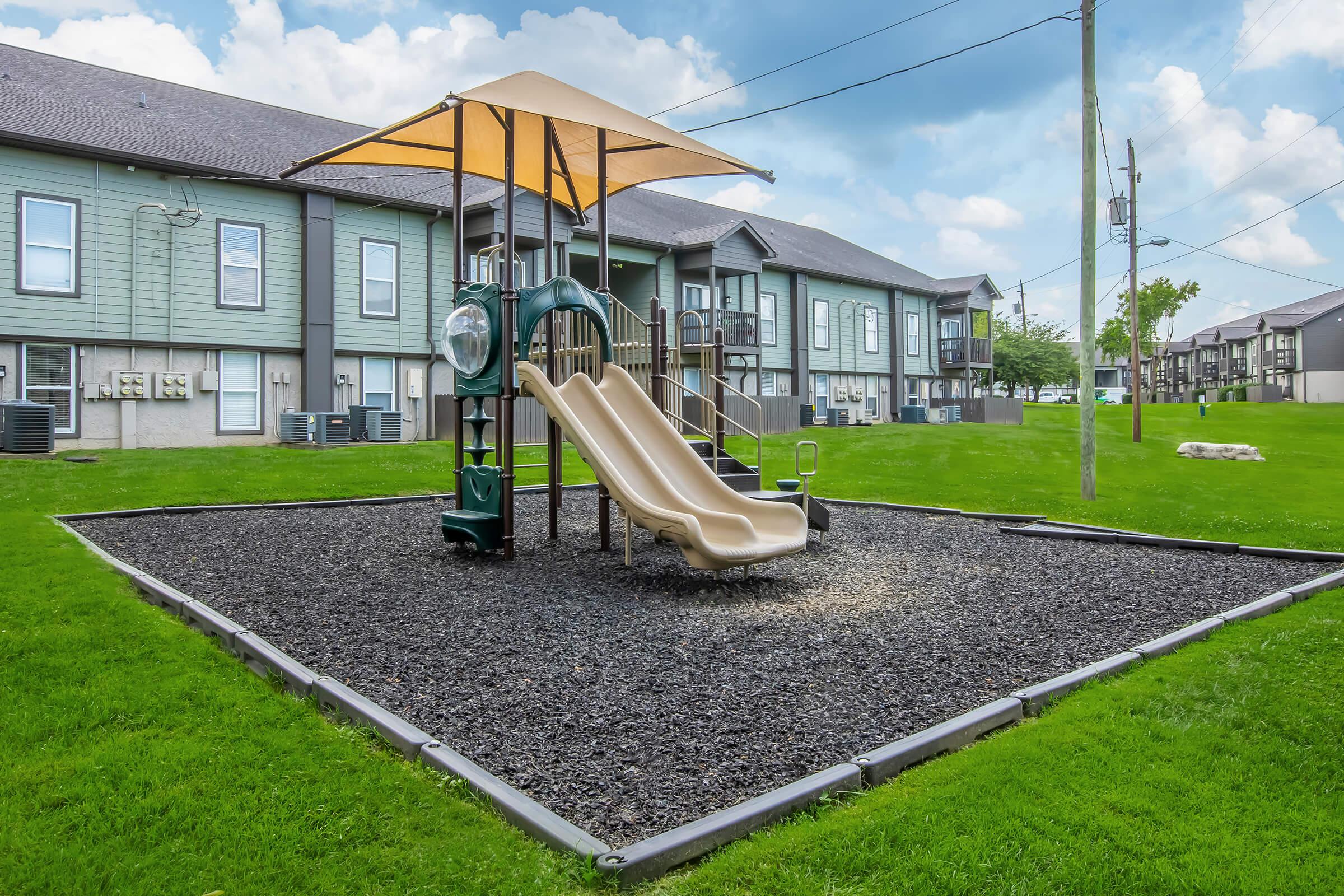 Playground area featuring two slides under a large canopy, surrounded by black rubber mulch. The background shows multiple residential buildings with green exteriors and manicured lawn. Overcast sky with scattered clouds above.