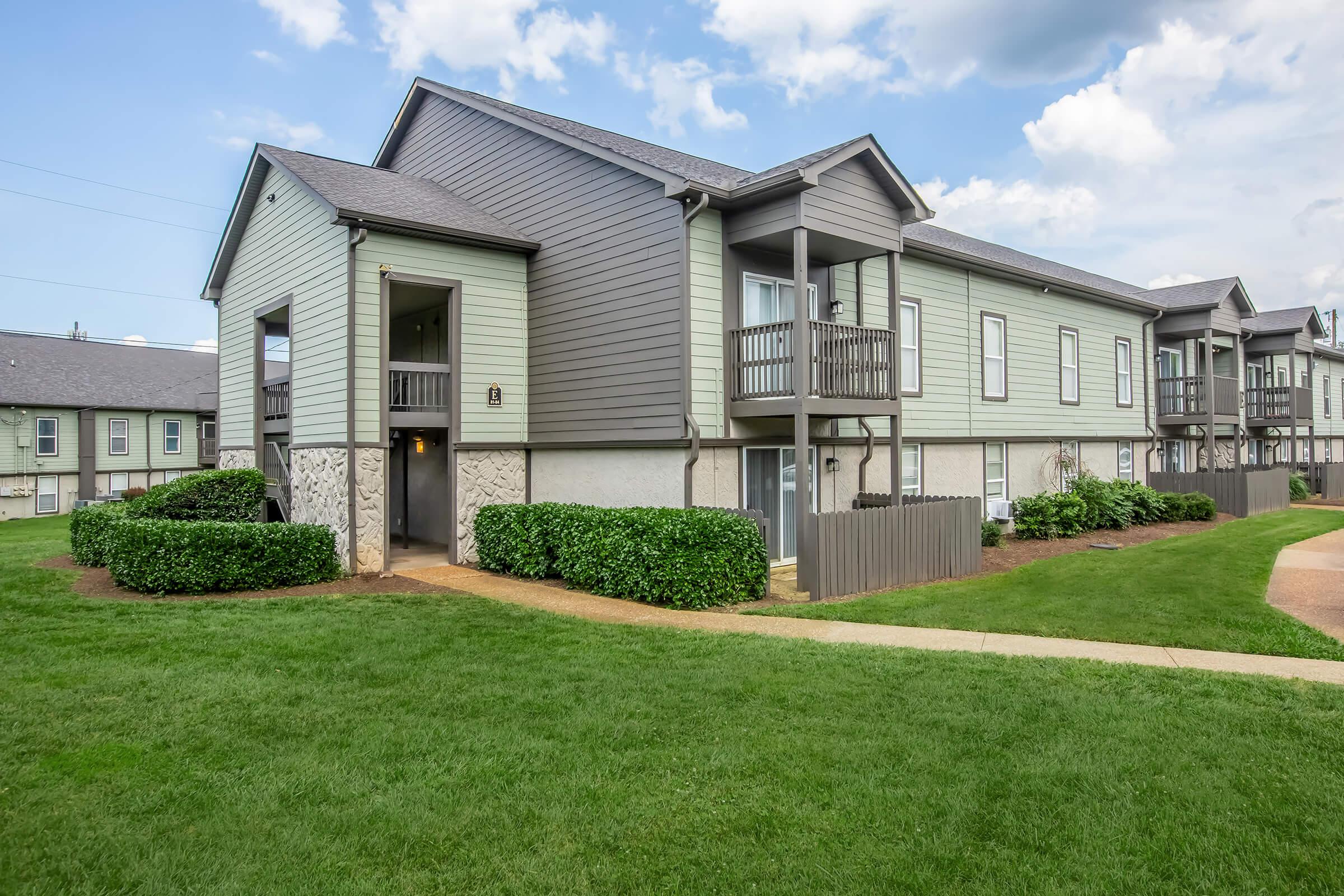 Two-story residential building with gray siding and balconies, surrounded by green lawns and shrubs. A pathway leads to the entrance, and a blue sky with clouds is visible overhead. The architecture features a mix of modern and traditional design elements.