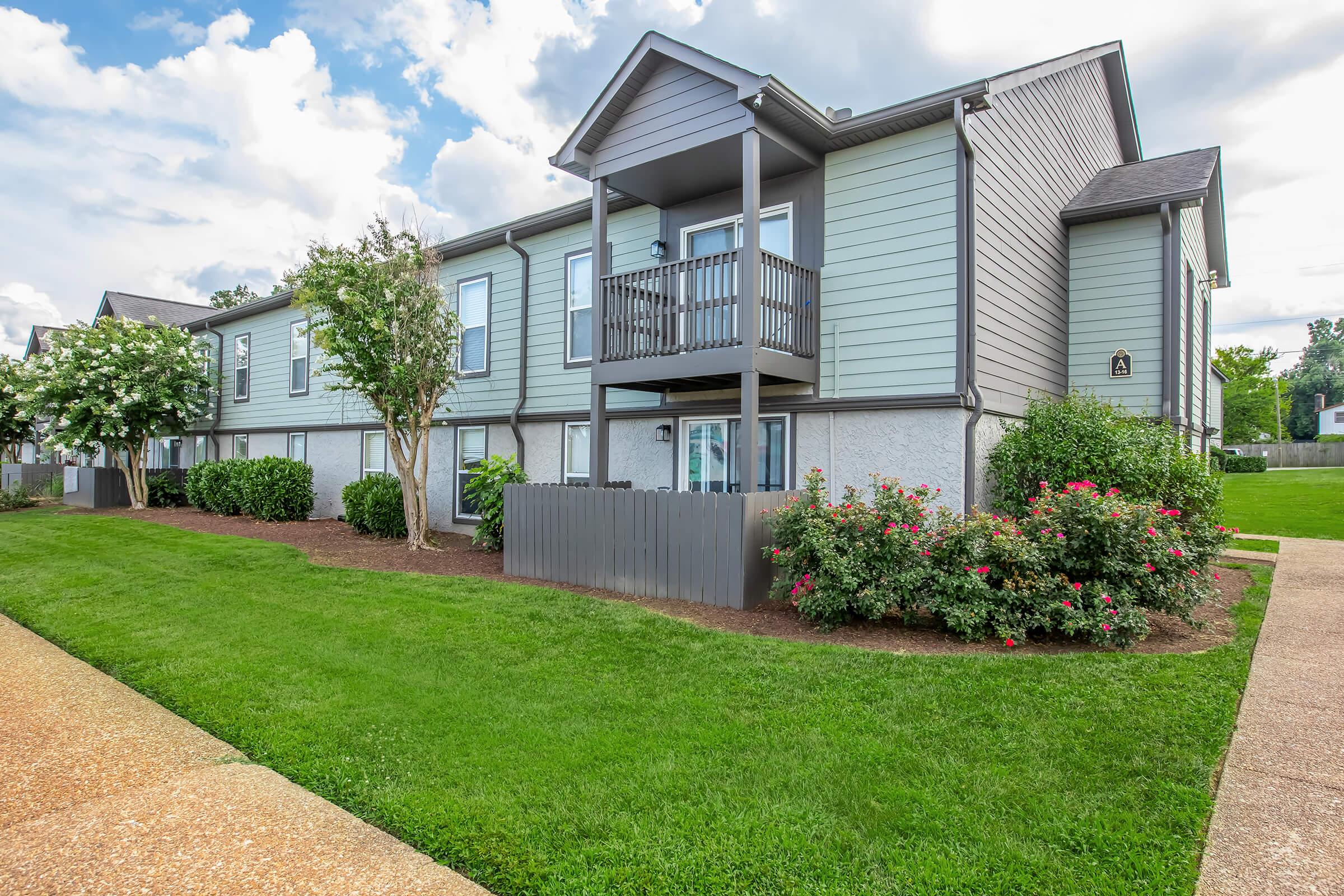 A two-story apartment building with light green siding, featuring balconies on the upper floor. The landscape includes well-maintained grass, shrubs, and flowering plants. The sky is partly cloudy, creating a pleasant, inviting atmosphere.