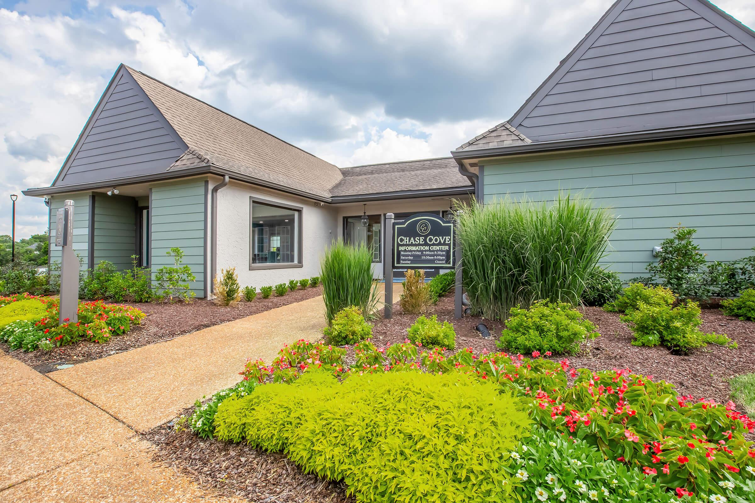 A modern building named "Chase Cove," featuring a welcoming entrance surrounded by well-maintained landscaping with vibrant flowers and greenery. The sky is partly cloudy, adding to the inviting atmosphere of the property.