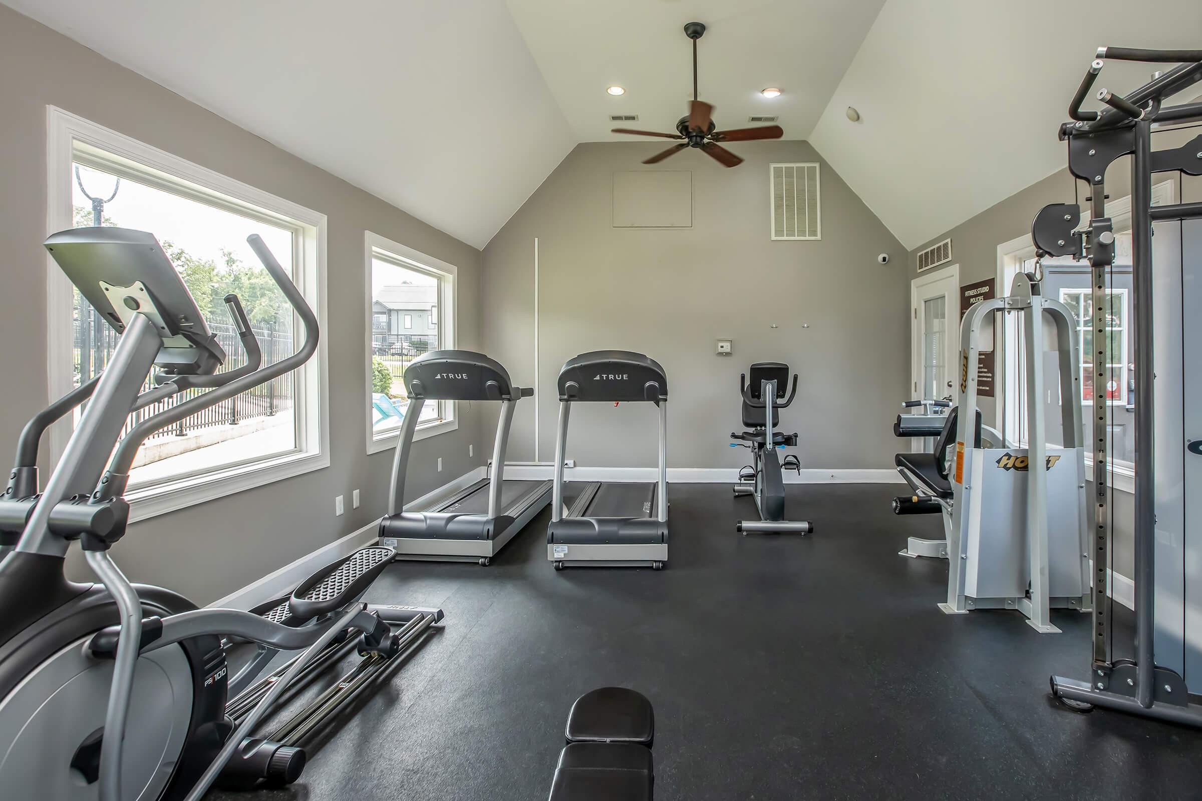Interior of a modern gym featuring various exercise equipment, including treadmills, an elliptical machine, weight machines, and free weights. The room has high ceilings, large windows allowing natural light, and a clean, organized layout.