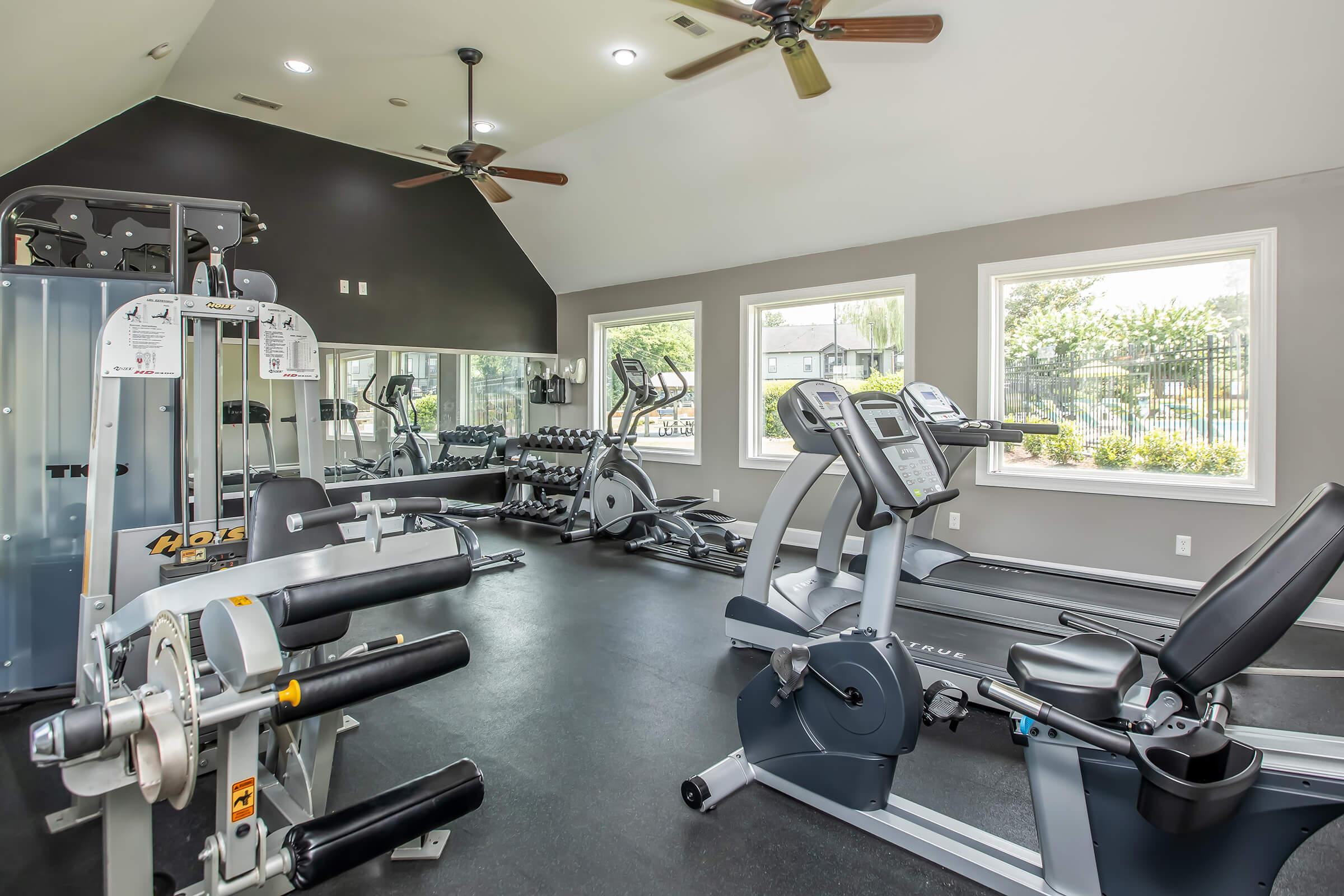 A modern gym interior featuring various workout equipment, including weight machines and exercise bikes. Large windows provide natural light, and a sleek design with dark walls and light flooring creates an inviting atmosphere. Ceiling fans enhance ventilation for a comfortable workout environment.