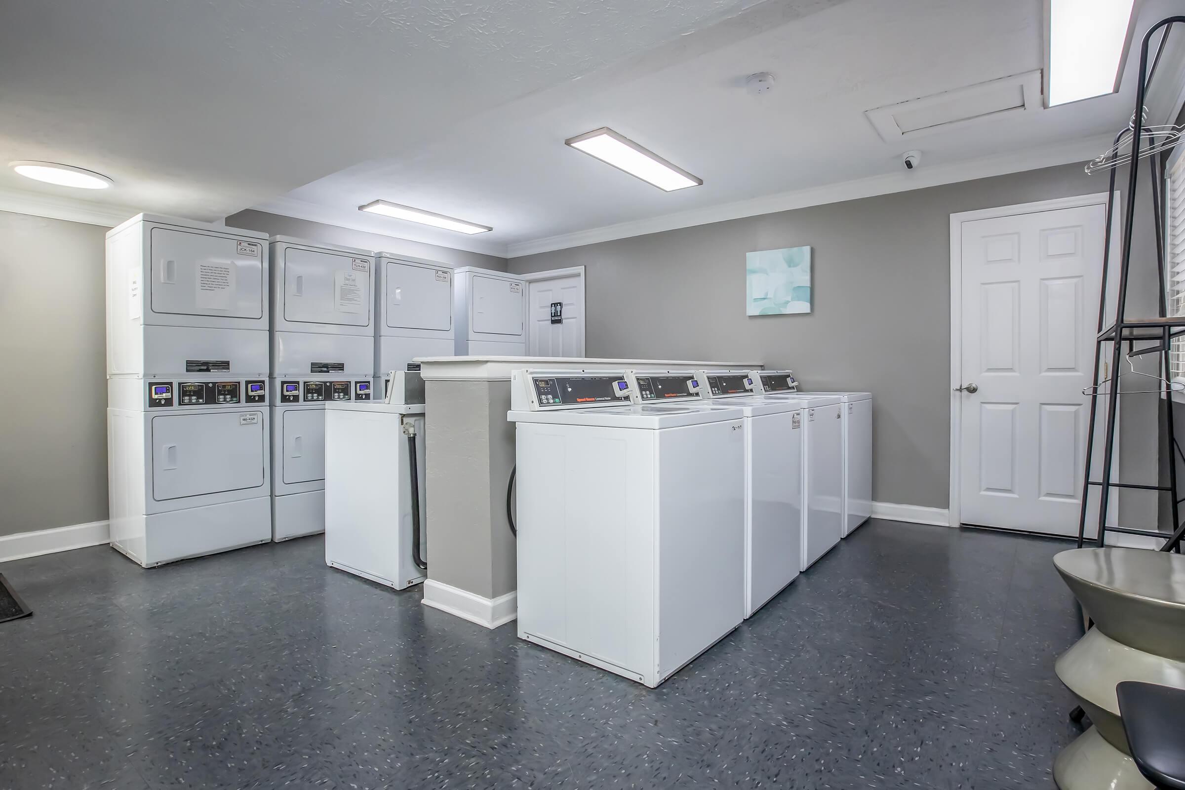 A clean and modern laundry room featuring several white washing machines and dryers arranged against a gray wall. The flooring is dark, and there are bright overhead lights. A door leads to another area, and there is a small shelf for folding clothes. The overall atmosphere is neat and well-maintained.