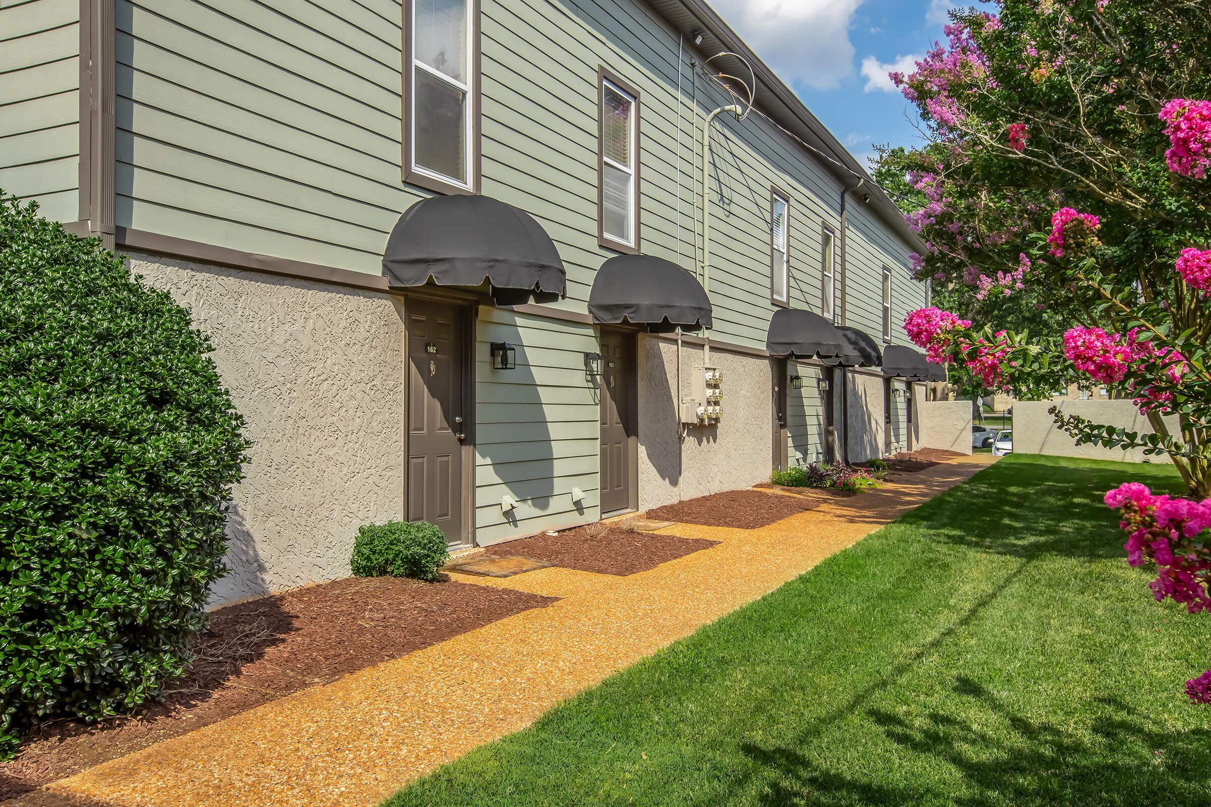 A pathway lined with purple flowering bushes leads to a series of green and gray apartment units, each with a black awning over the entrance. The well-maintained lawn features mulch and landscaping, creating a tidy residential environment.