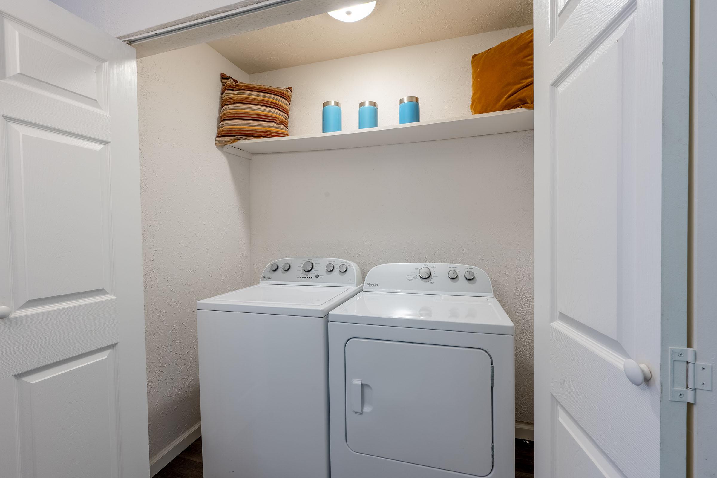 A laundry nook featuring a white washing machine and dryer side by side. Above them, a shelf holds three blue canisters and a brown pillow. The space is well-lit and enclosed by white doors, creating a tidy and organized laundry area.