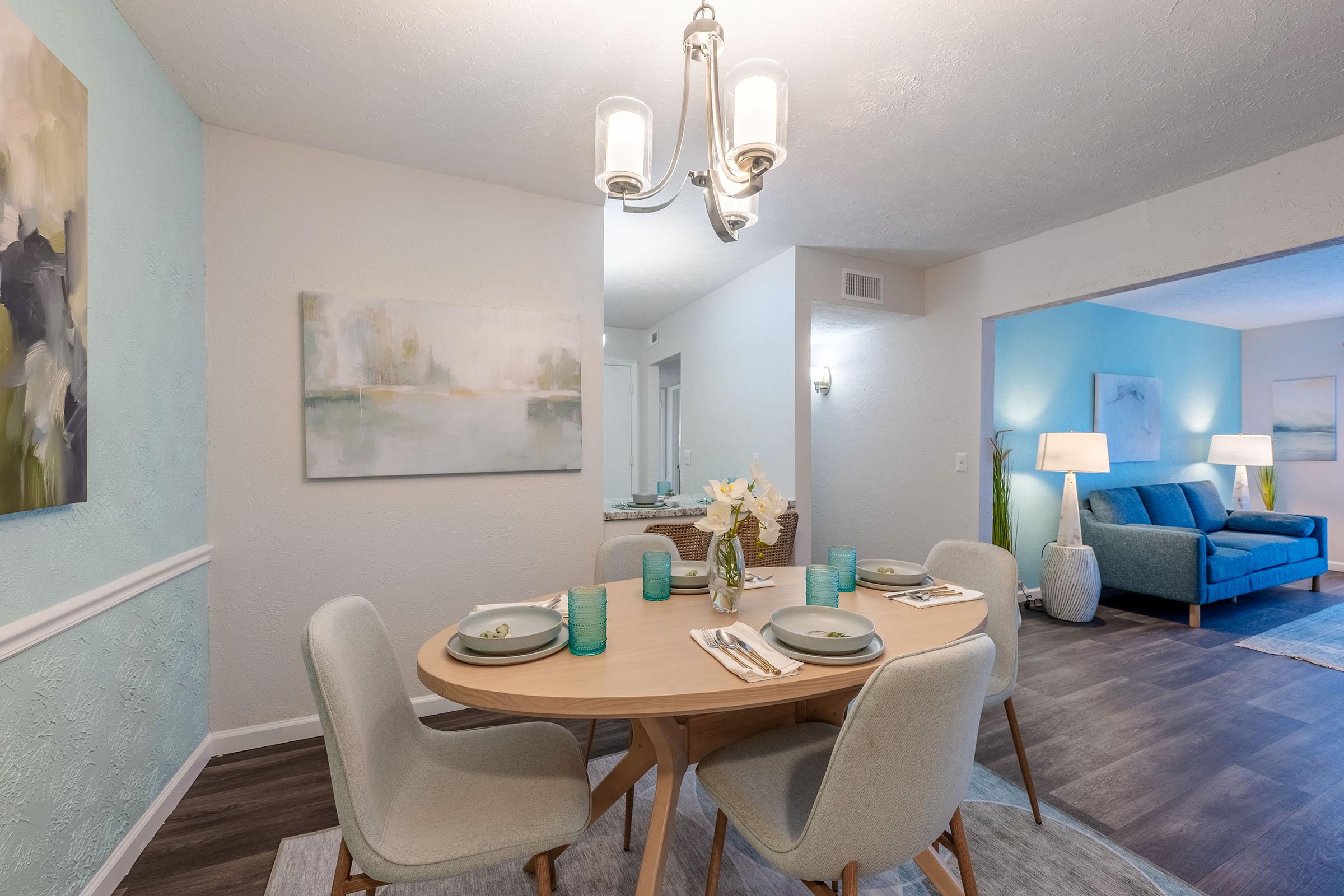 A bright dining area featuring a round wooden table set with light grey chairs, turquoise glasses, and a vase of flowers. Soft lighting is provided by a stylish chandelier. The walls are painted in light colors, with abstract artwork displayed. A cozy living area with a blue couch can be seen in the background.