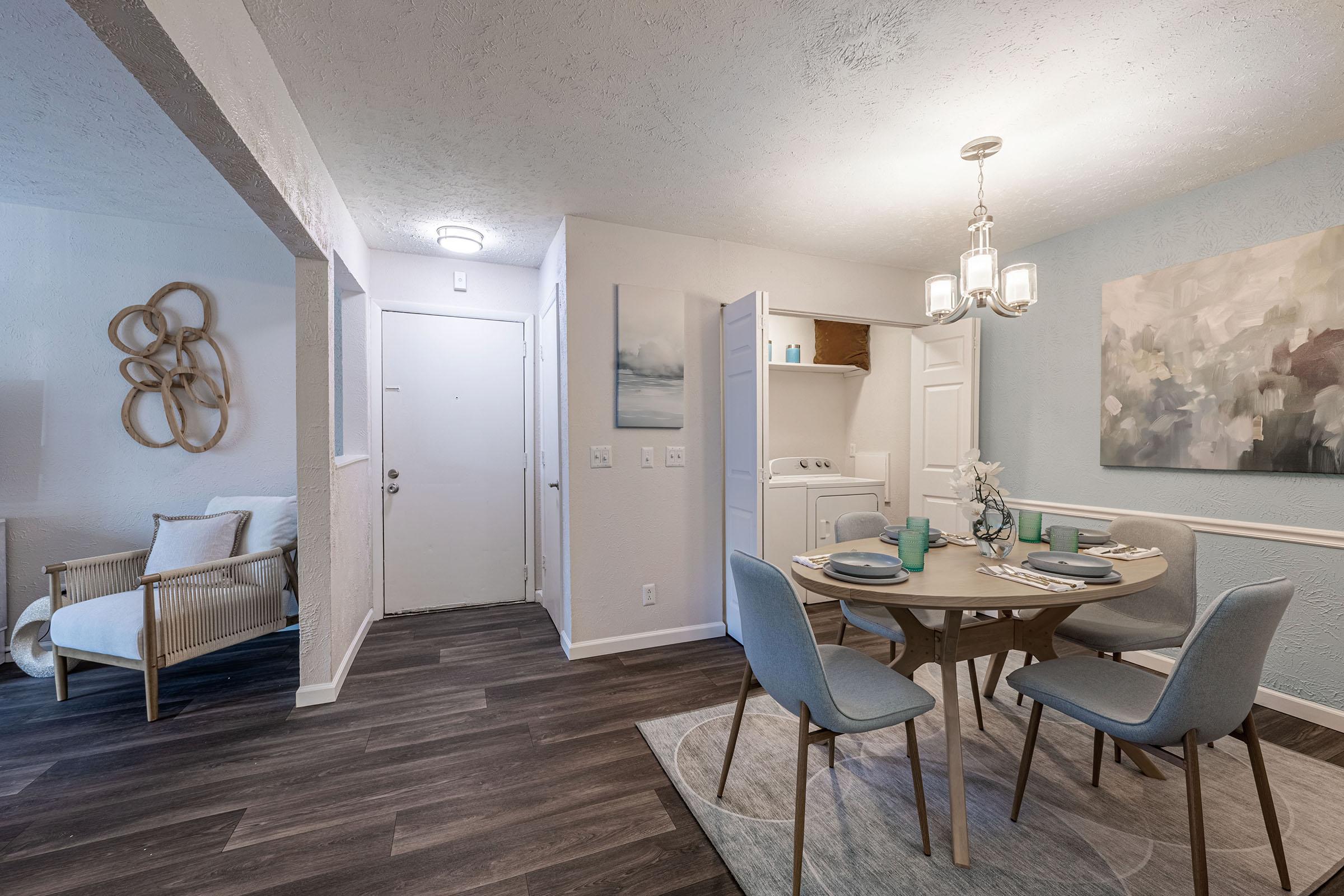 A modern dining area featuring a round wooden table with light blue table settings, surrounded by four light gray chairs. A cozy armchair is nearby, with a decorative wall piece and soft lighting creating a welcoming atmosphere. The entrance door is visible, along with a partially open kitchen area.