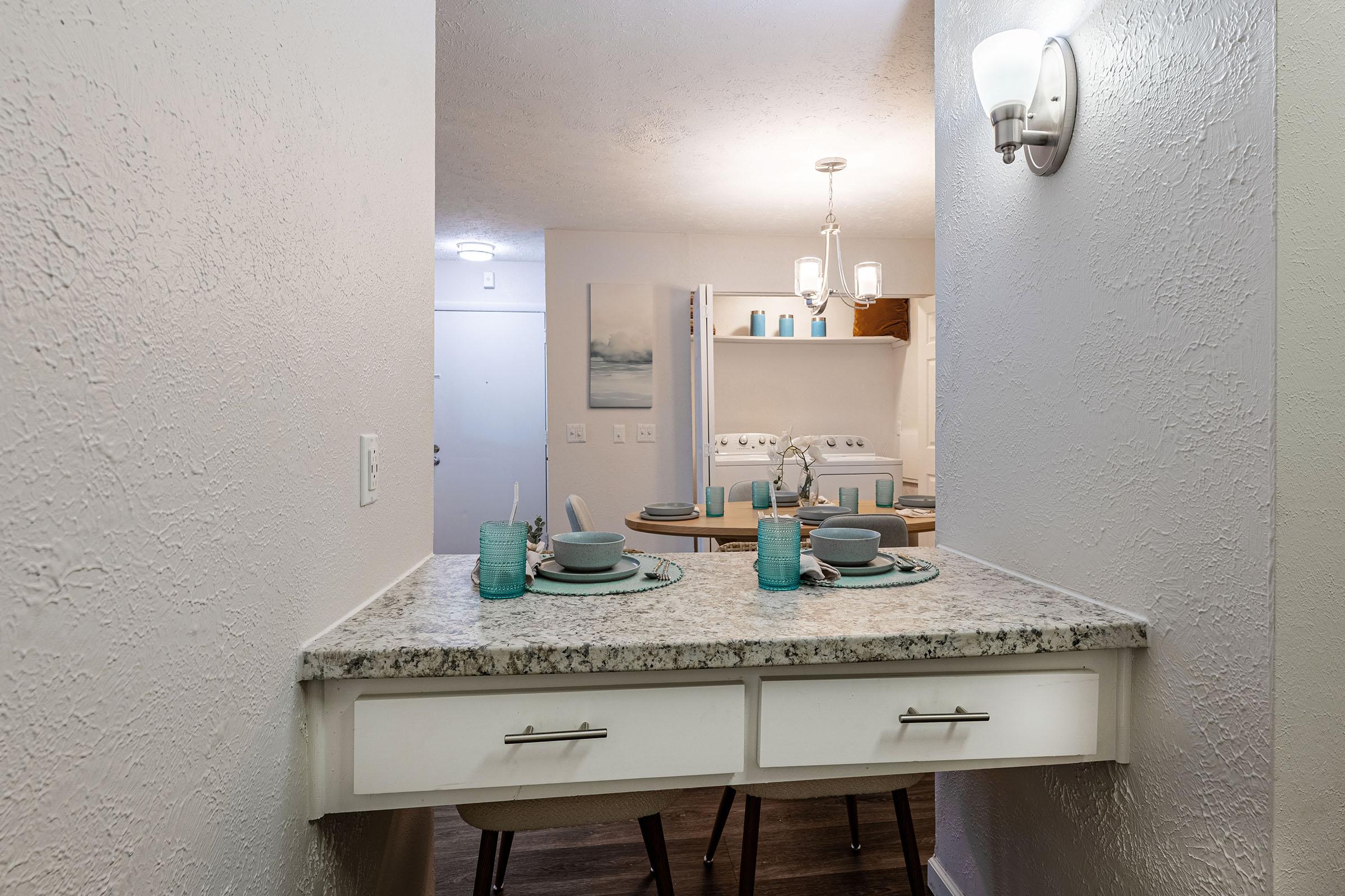 Modern kitchen view from a hallway, featuring a granite countertop with table settings including plates, cups, and utensils. Light-colored walls and a pendant light above enhance the bright atmosphere. The dining area is visible in the background, showcasing additional decor and tableware.