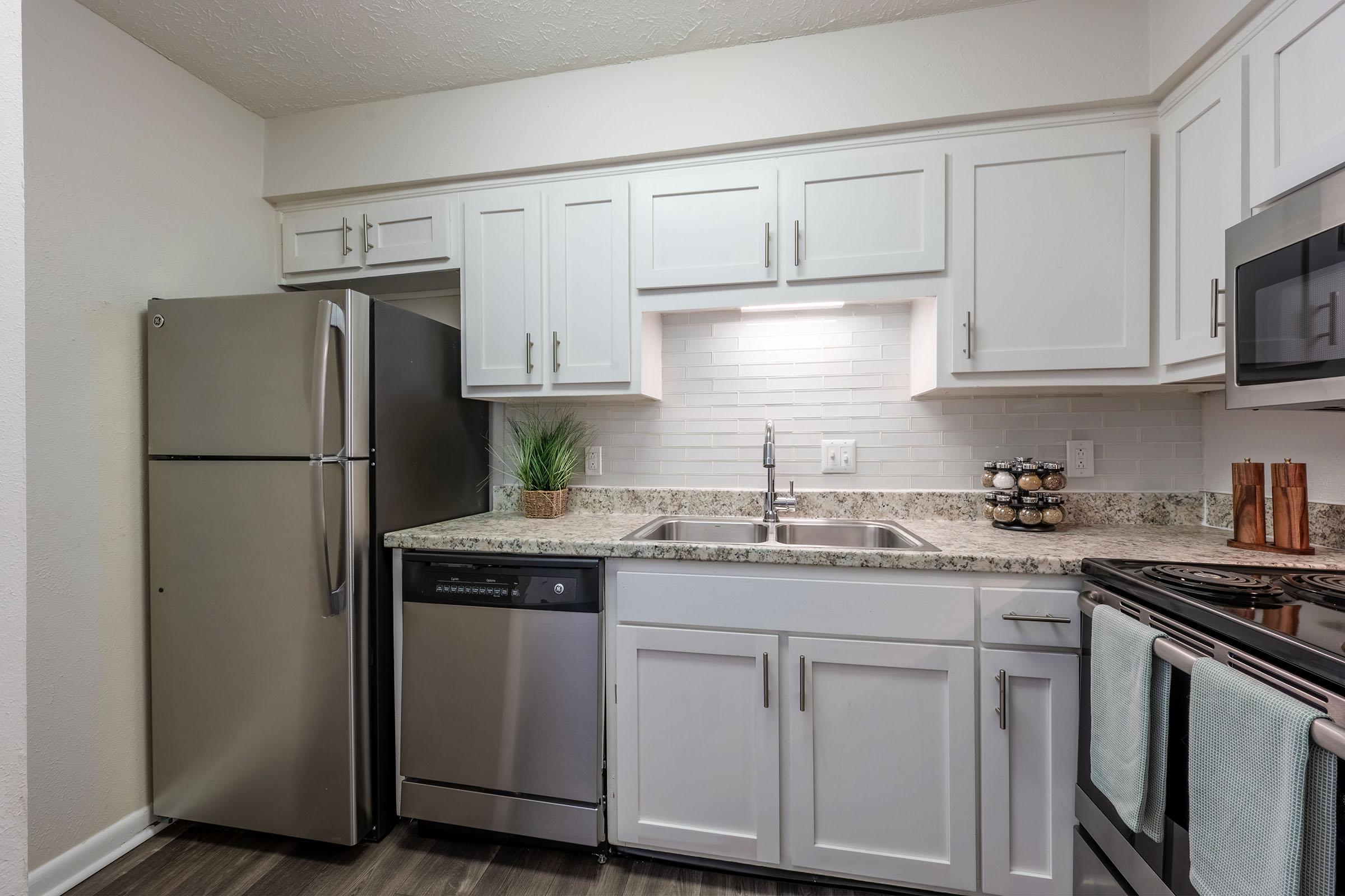 A modern kitchen featuring white cabinetry, a stainless steel refrigerator, a dishwasher, and an oven. The countertop is made of granite, with a backsplash of white tiles. There is a small plant near the sink and some decorative items on the counter. The flooring is a dark wood laminate.