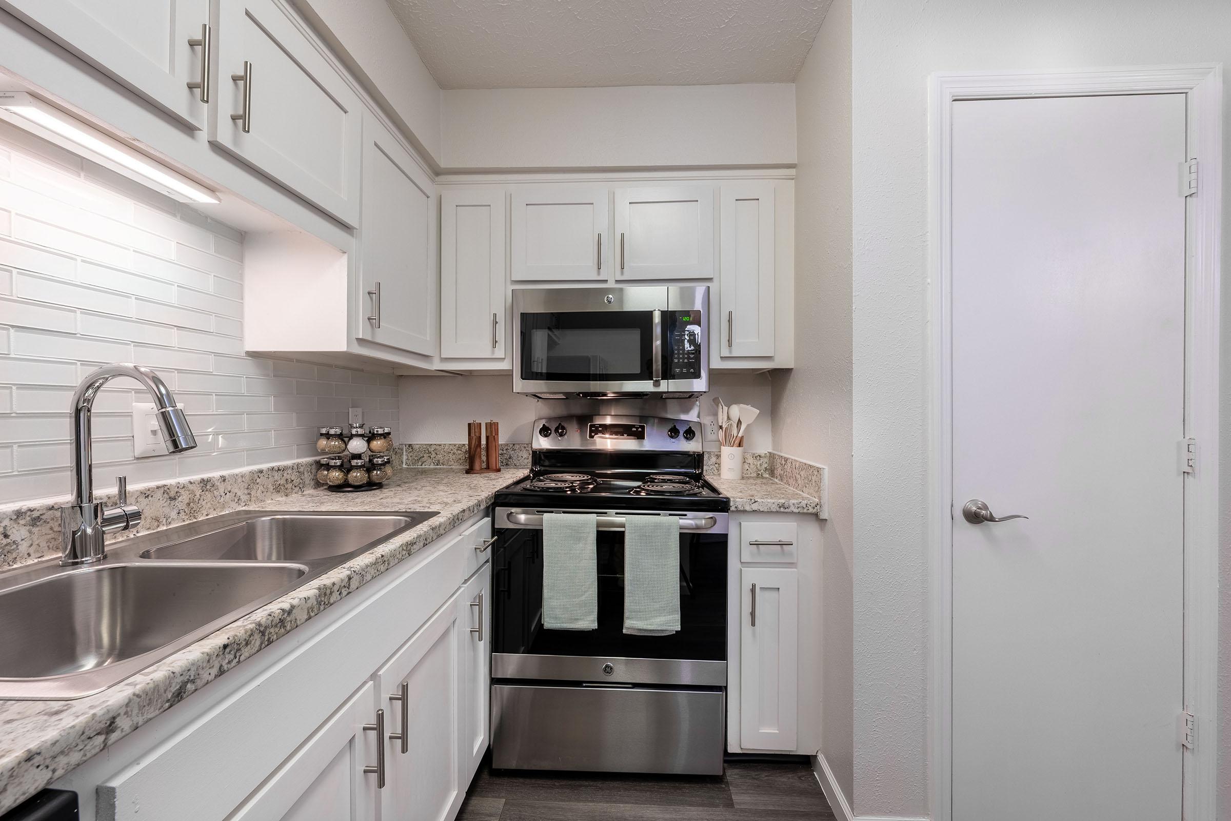 A modern kitchen with white cabinets, a stainless steel oven, and a microwave above it. The countertops are made of light granite, and a double sink is visible. Kitchen towels hang from the oven. There are jars on the countertop and a white door on the right leading to another room.