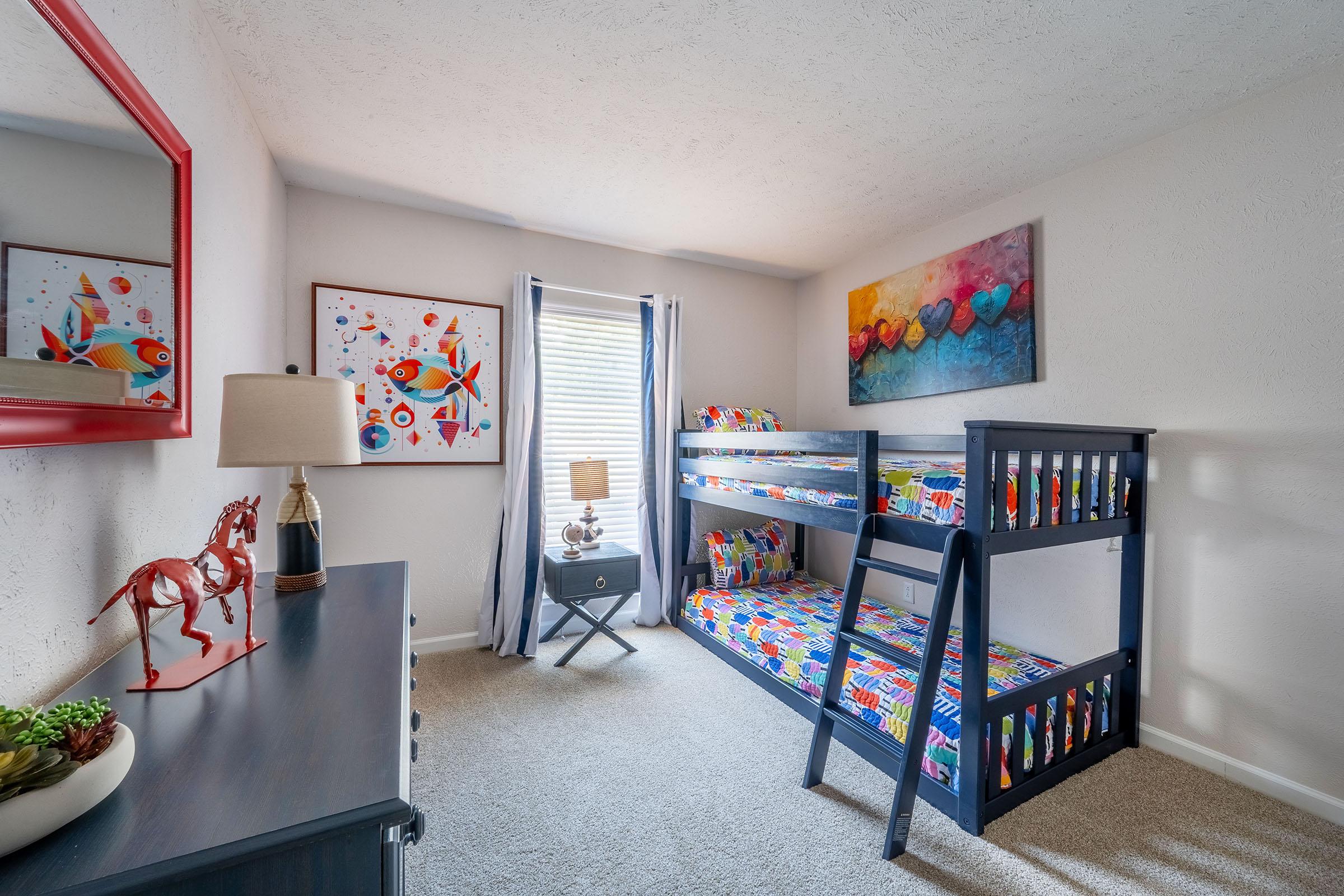 A colorful child’s bedroom featuring a bunk bed with vibrant bedding, a small nightstand with a lamp, a dresser, and decorative artwork on the walls. Natural light streams through a window with curtains, creating a cheerful and inviting atmosphere. A decorative horse figurine and some plants add charm to the space.