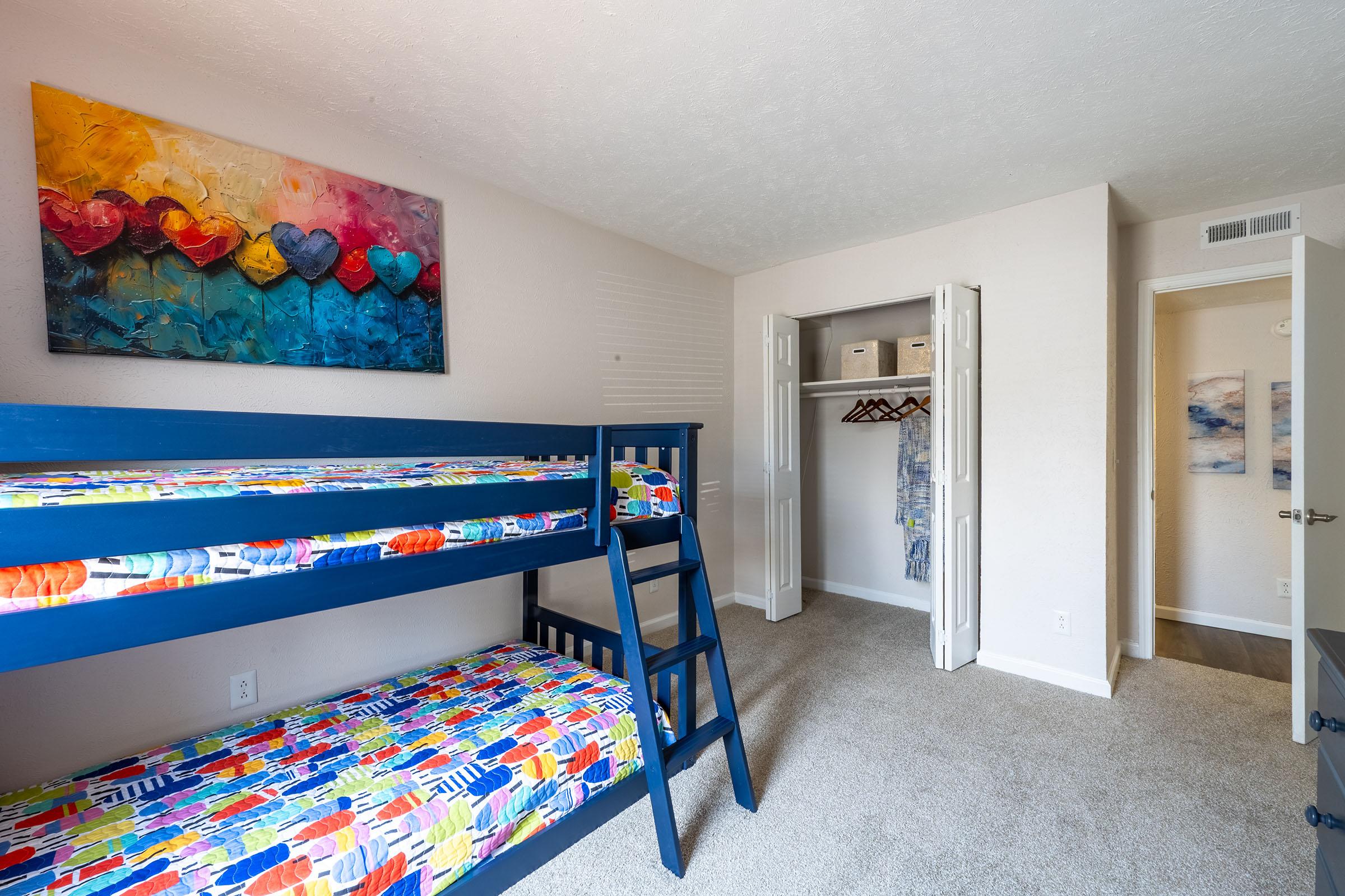 A bright and colorful children's bedroom featuring a blue bunk bed with patterned bedding. A colorful abstract painting with hearts hangs above the bed. The room has beige walls and carpet, and a closet with sliding doors is visible, showcasing hangers and organized clothes. Natural light illuminates the space.