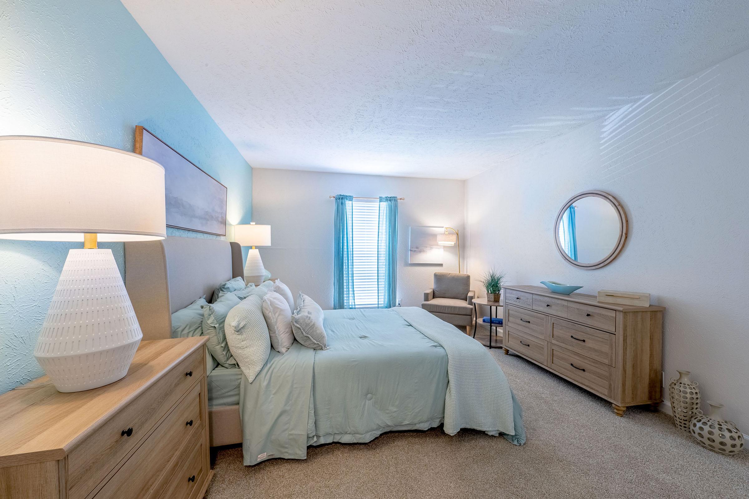 A bright, airy bedroom featuring a light blue accent wall, a queen-sized bed with soft turquoise bedding, matching lamps on bedside tables, and a wooden dresser. A cozy armchair sits near a window, which allows natural light to fill the space, enhancing the serene atmosphere.