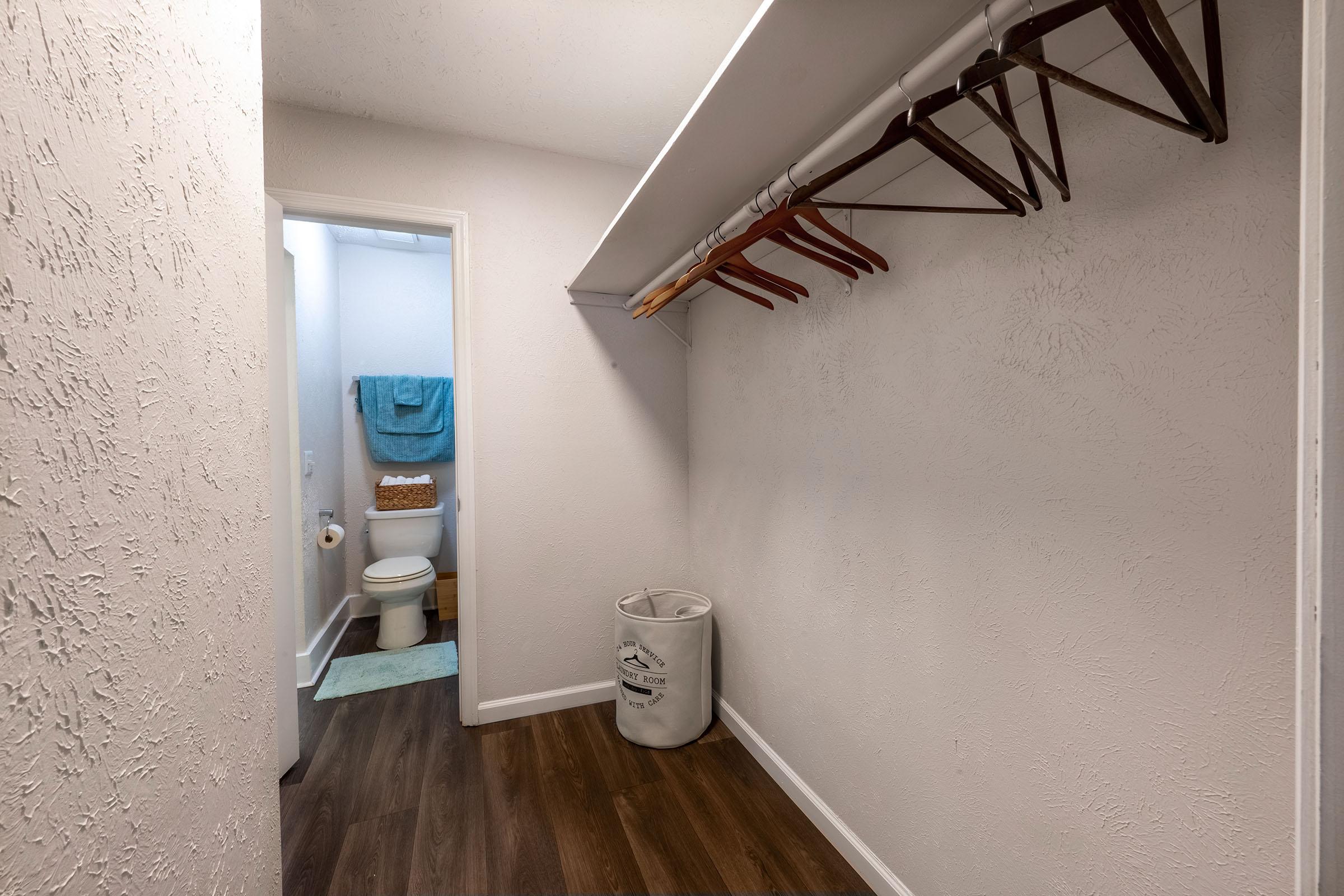 A hallway leading to a bathroom, featuring a closet area with wooden hangers on a rod, a white laundry basket on the floor, and light-colored walls. The bathroom door is partially visible at the end of the hallway, with a teal towel hanging on the door. The floor is dark wood laminate, adding warmth to the space.