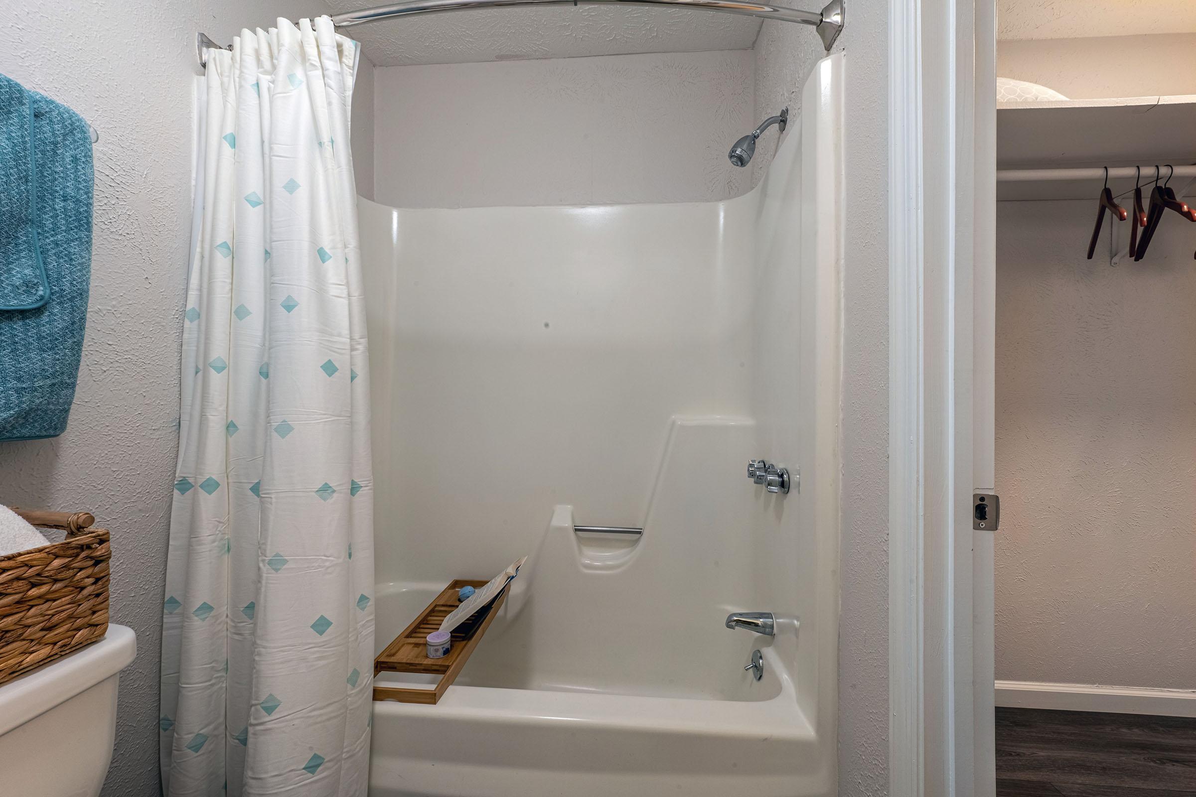 A clean, light-colored shower with a glass door partially open, featuring a white shower curtain with blue diamonds. A showerhead is mounted on the wall, and a wooden bath caddy is resting on the edge. A basket with towels is nearby, and a closet with hanging items is visible in the background.
