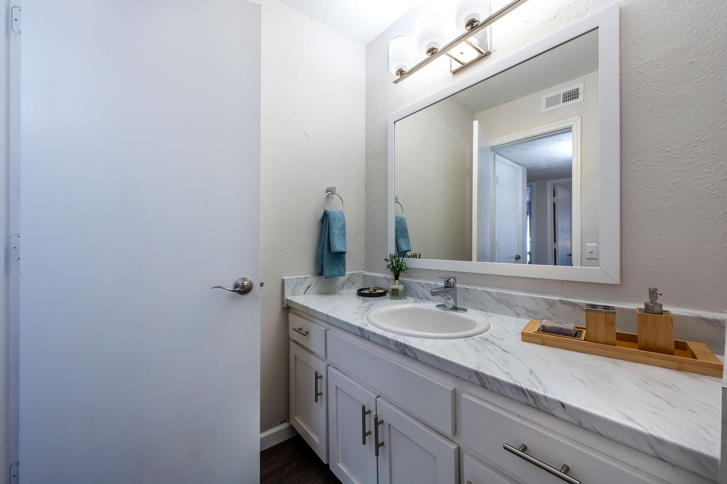 A clean bathroom featuring a white vanity with a marble countertop, a round sink, and a large mirror above. There are two blue towels hanging on the wall, a small potted plant on the counter, and a wooden tray with bathroom accessories. A door leads to another room in the background.