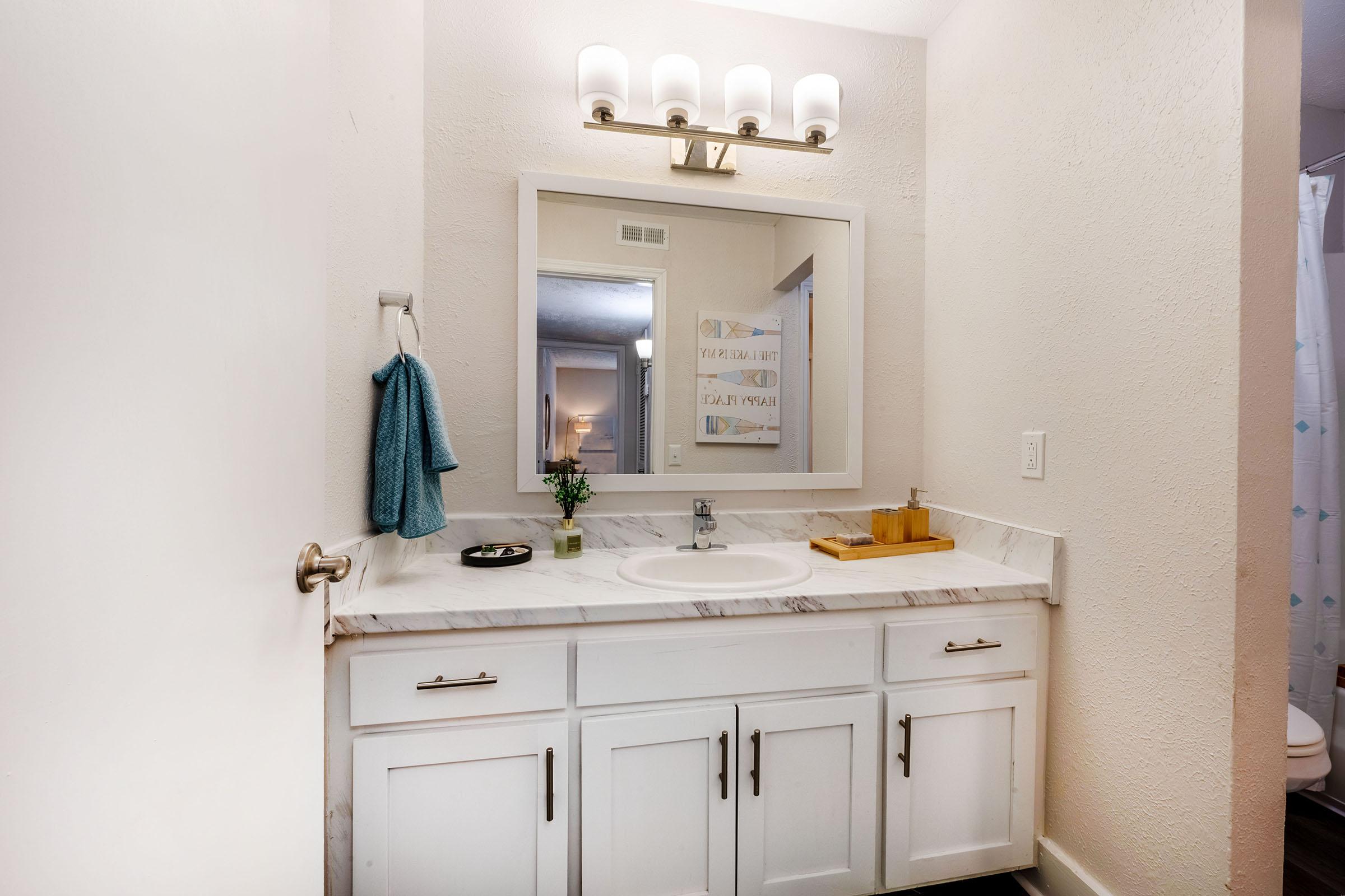A bathroom with white cabinetry, a marble countertop, and a sink. There's a large mirror above the sink, a light fixture with three bulbs, and a blue hand towel hanging. A small potted plant and a wooden tray are on the counter. In the background, a shower curtain is visible. The walls are painted light and the space is well-lit.