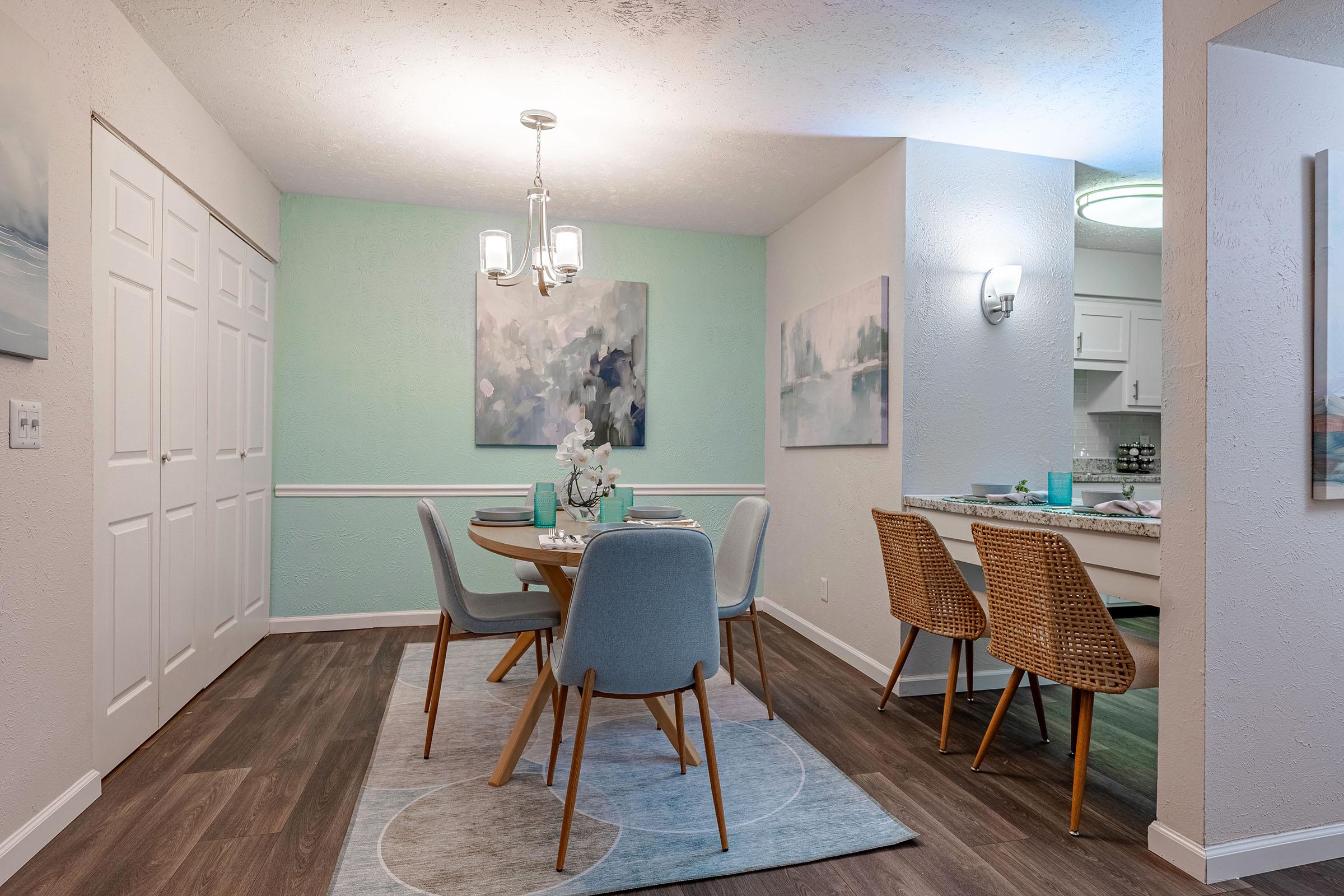 A modern dining area featuring a round table with light blue chairs, a decorative centerpiece, and a rug. The walls are painted in soft green and white, adorned with abstract artwork. Nearby is a kitchen area with light cabinetry, and woven chairs are positioned at a small adjoining table. Natural light fills the space.