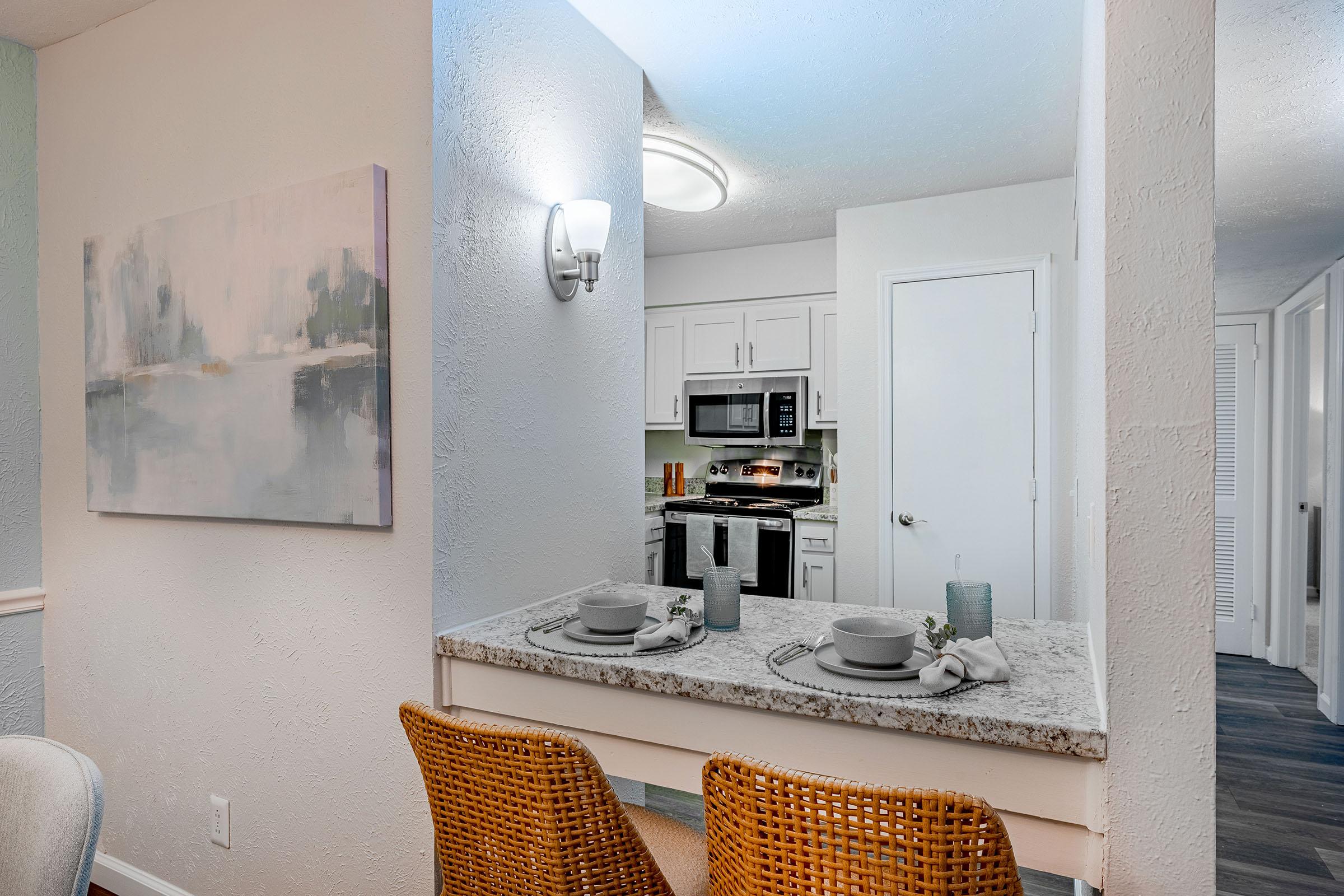 A modern kitchen with stainless steel appliances, including a microwave, oven, and refrigerator. The dining area features a granite countertop with two wicker bar stools and neatly set dishes. Soft lighting highlights a decorative painting on the wall, creating a cozy atmosphere.