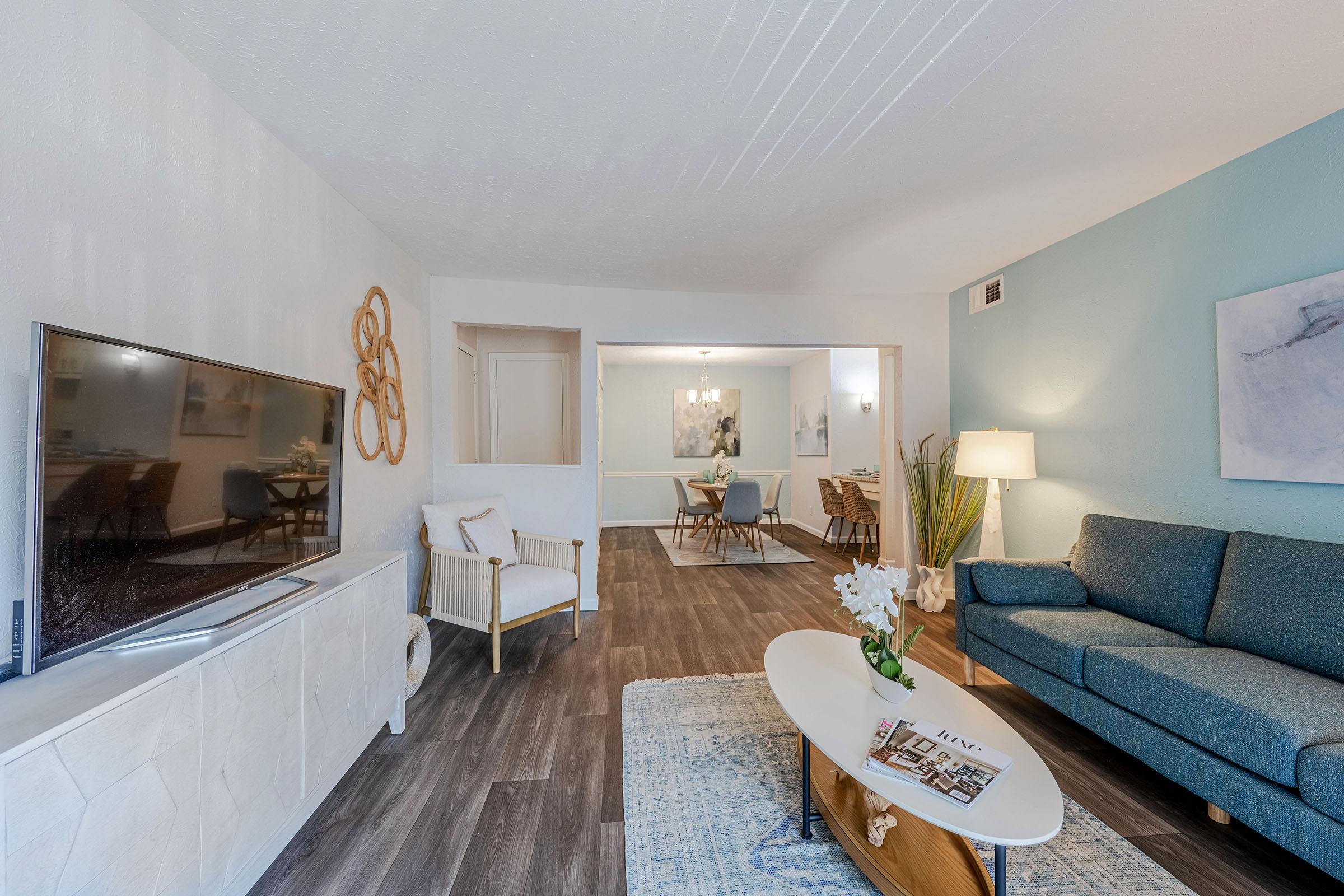 A modern living room featuring a comfortable blue sofa, a stylish coffee table, and a large flat-screen TV. The room is decorated with light-colored walls, a soft rug, and artistic wall decor. In the background, a dining area is visible with a table and chairs, illuminated by a ceiling light.