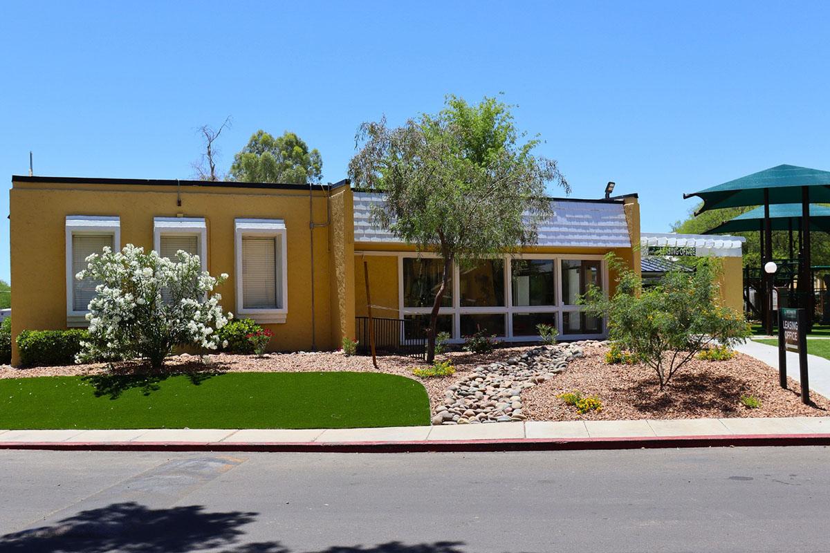 A single-story yellow building with a sloped roof, surrounded by green lawns and flowering shrubs. There are rocks placed in the landscaping, and a green awning nearby, under clear blue skies. The setting looks well-maintained and inviting.