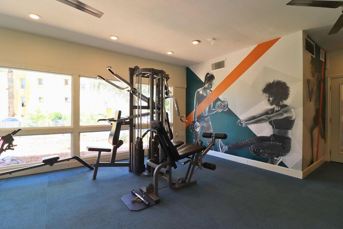 A modern fitness gym featuring a multi-station workout machine and a wall mural depicting athletes engaged in strength training. Large windows provide natural light and a view of the outdoors, creating an inviting exercise environment. The flooring is carpeted, adding comfort to the space.