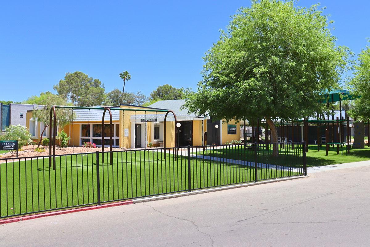 A view of a school or community center featuring a green lawn enclosed by a black fence. The building is yellow with several windows, surrounded by trees and desert landscaping under a clear blue sky. A playground structure is visible in the background, suggesting a family-friendly environment.
